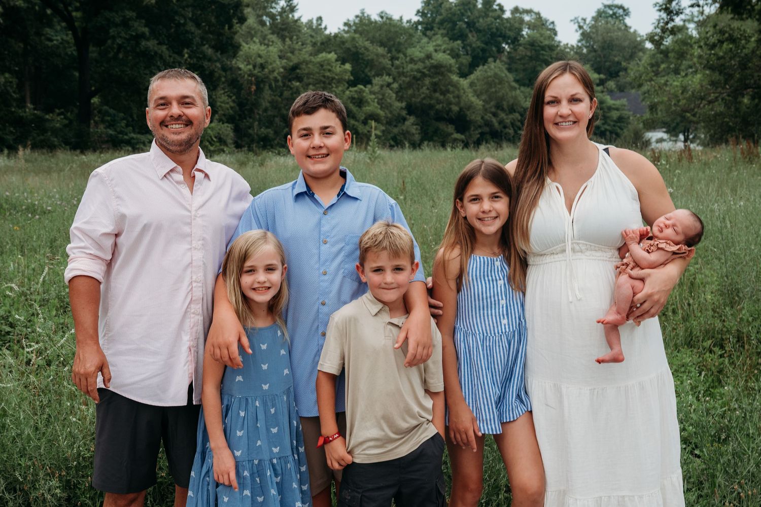 A family of seven poses together outdoors in a grassy field surrounded by trees on a summer day.