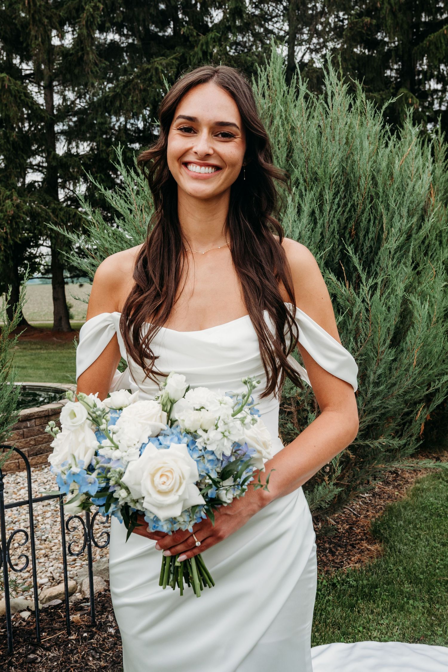 Smiling bride in off-shoulder white gown holding blue and white floral bouquet outdoors among evergreen trees.