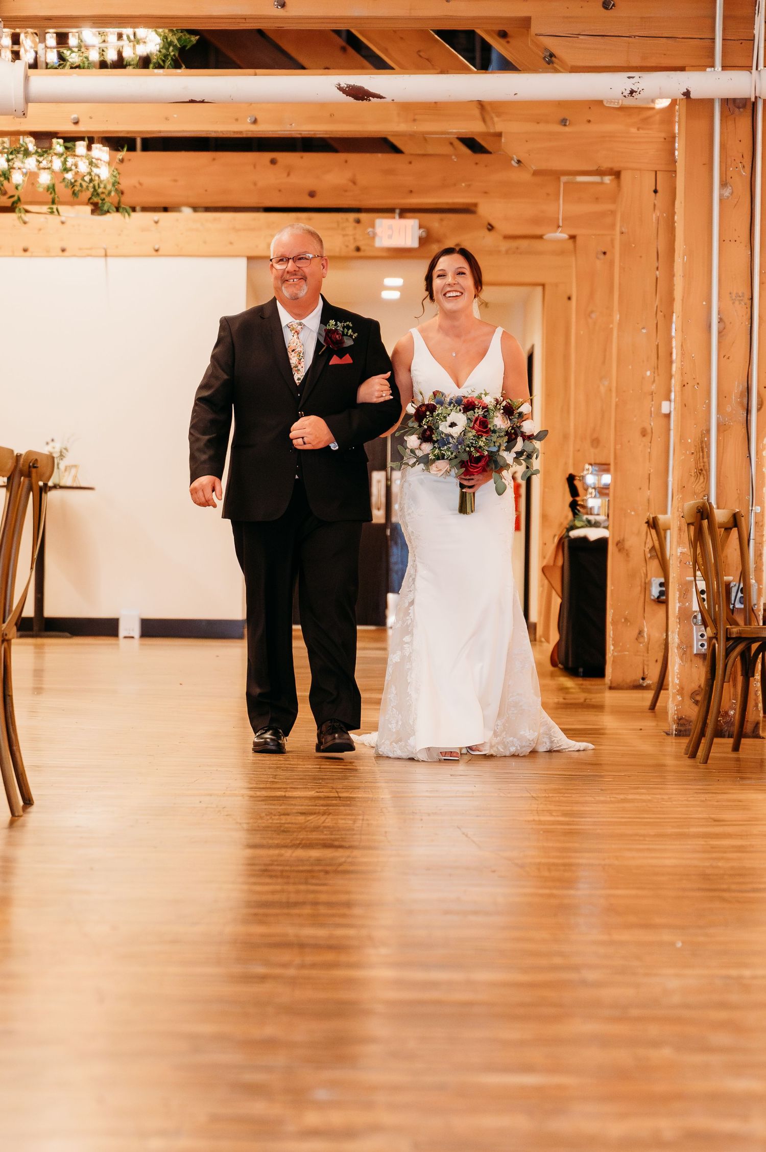 Wedding party walks down a wooden-floored aisle in a rustic barn venue with warm lighting and natural wood beams.