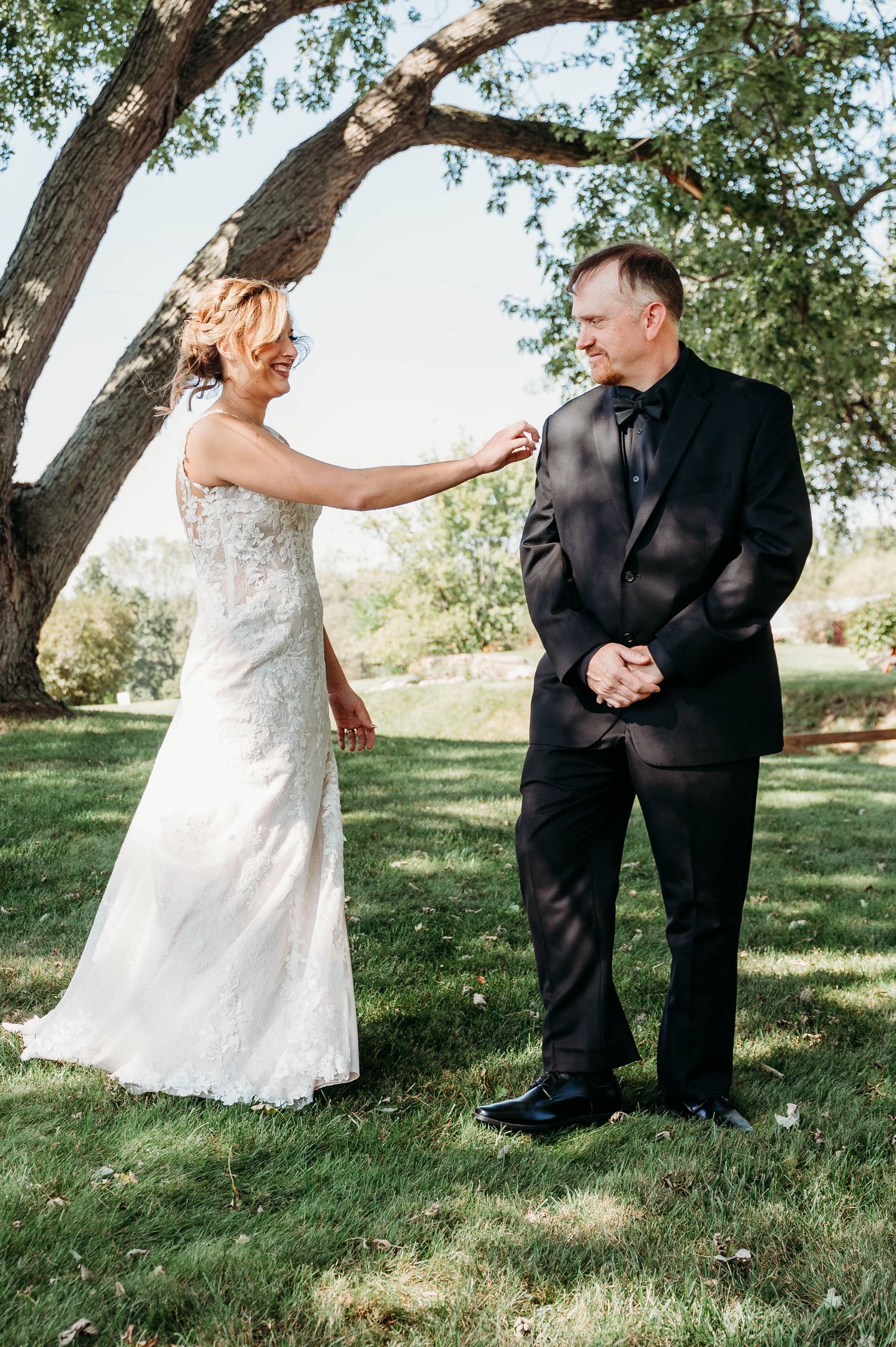 A couple in wedding attire shares playful moments under trees in a grassy outdoor setting.