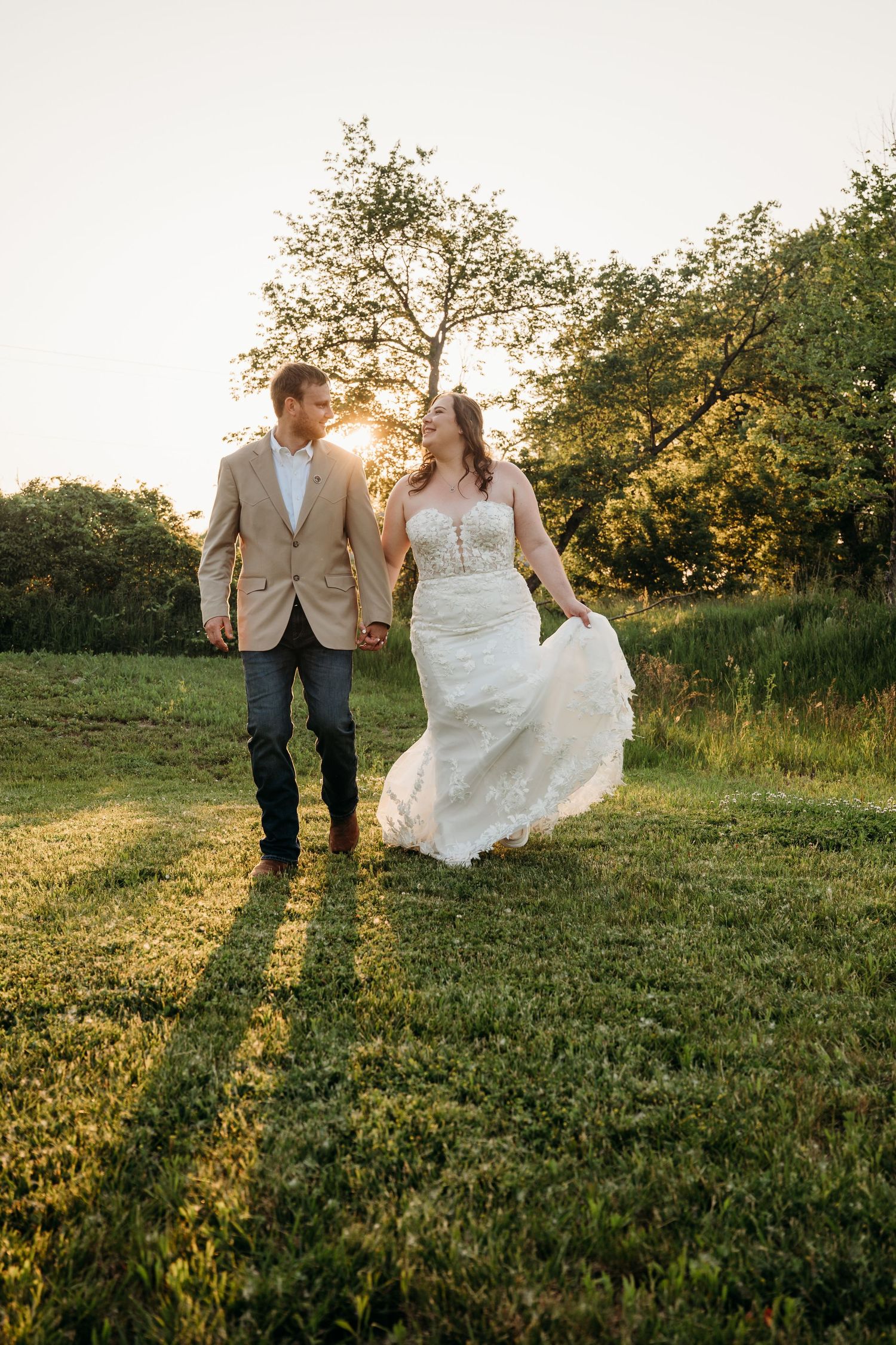 A couple walks hand in hand through a sunlit meadow at golden hour, with trees silhouetted against the setting sun.