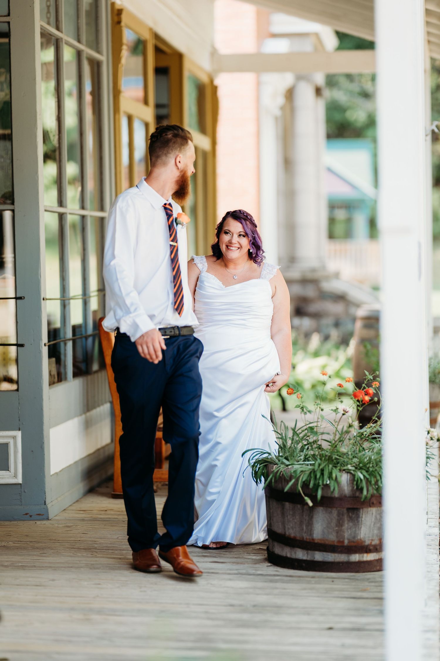 Romantic wedding couple sits together on a covered porch with rustic wooden barrels and potted plants.