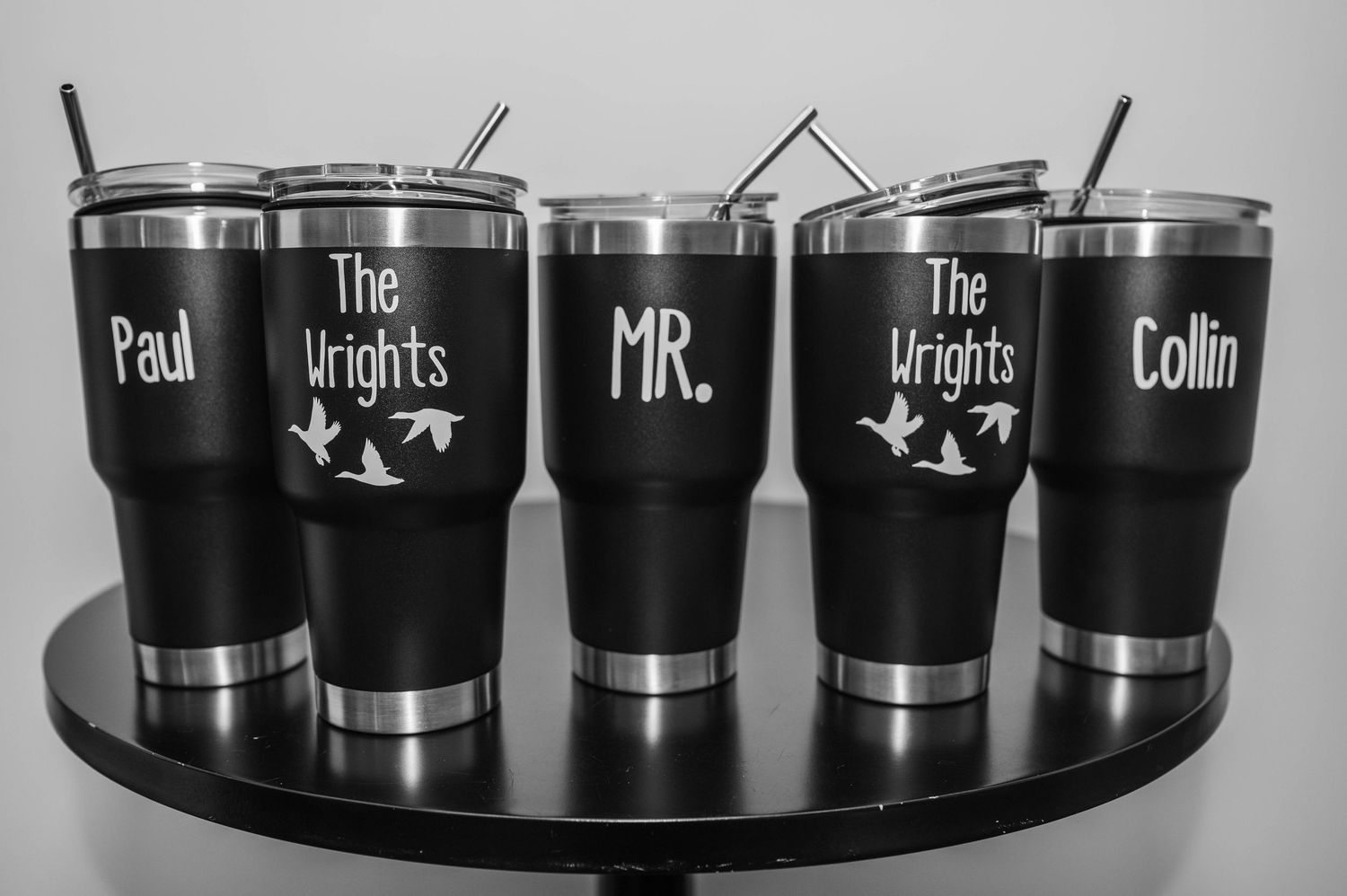 Black metal tumblers with band names engraved in white text displayed on a wooden surface.