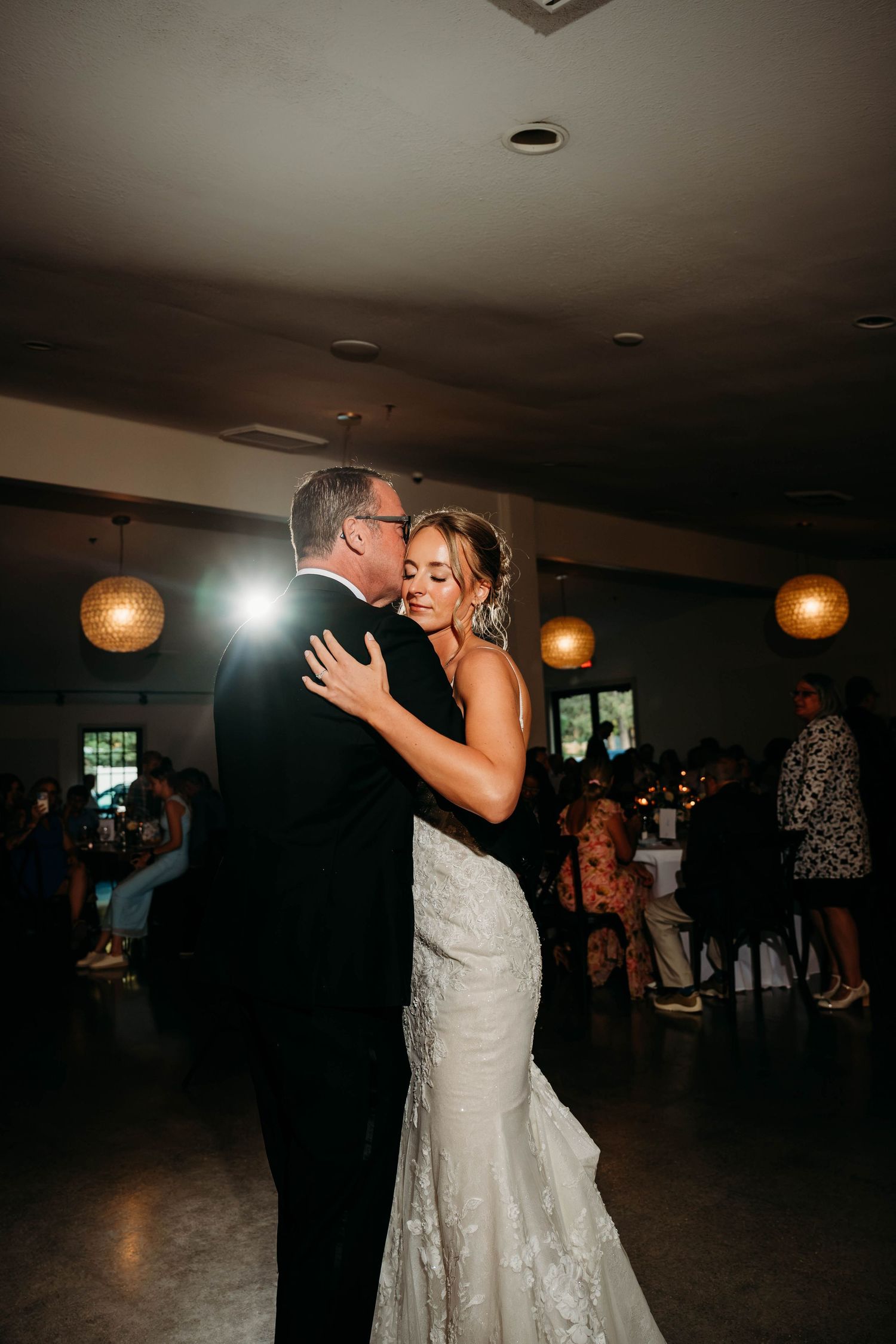 Bride and father share an emotional first dance at elegant wedding reception with warm pendant lighting.