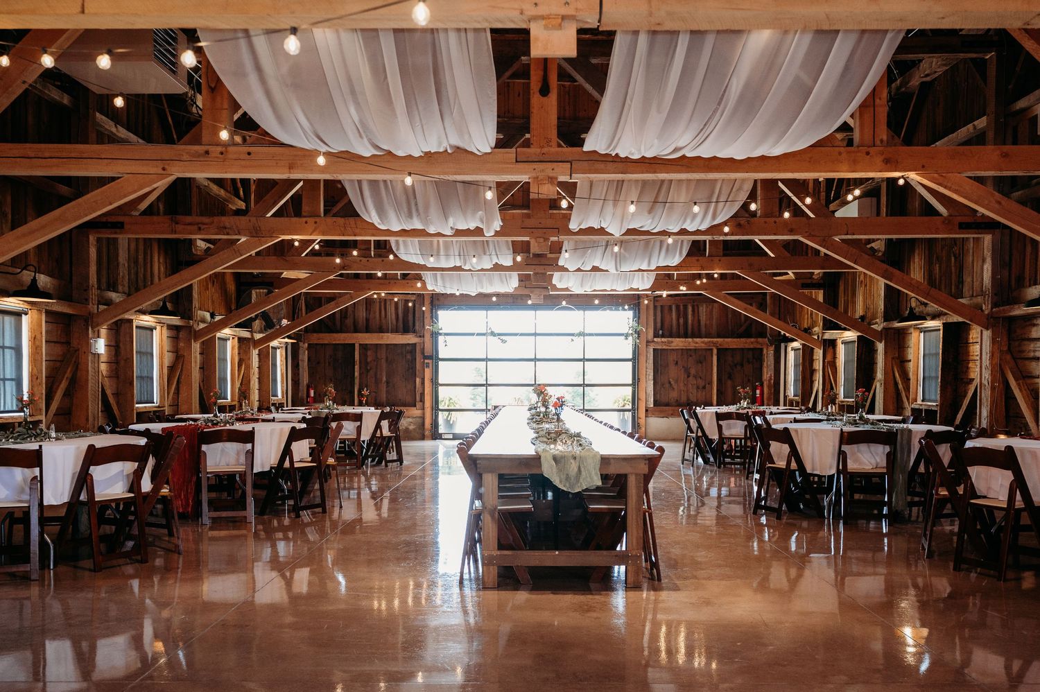 Large rustic barn venue with white draping and elegant lighting setup for a wedding reception.