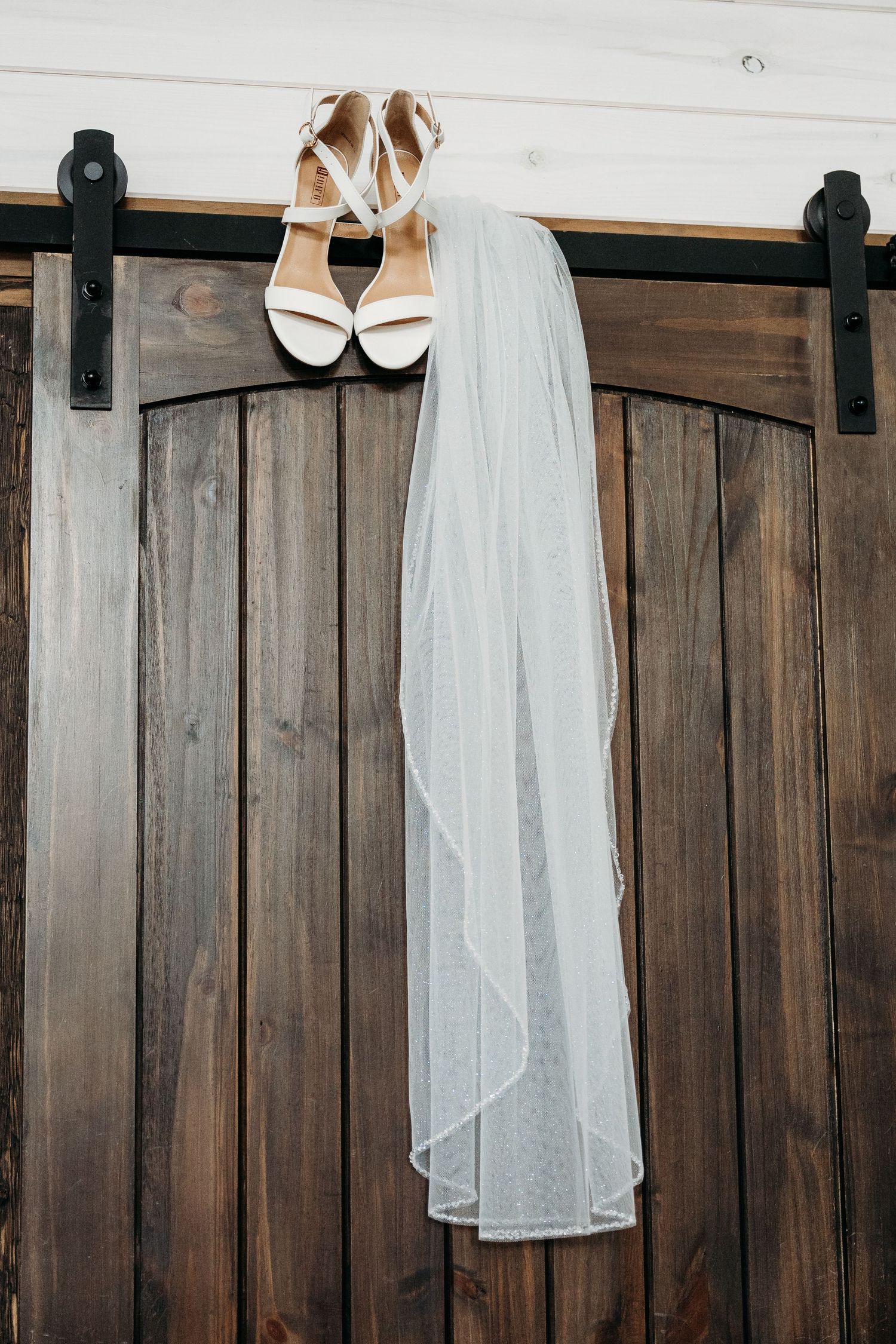A white wedding dress, veil, and shoes hang against a rustic wooden barn door with black hardware.