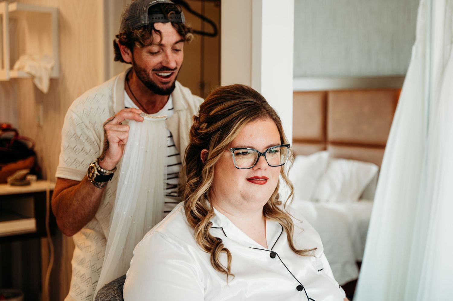 A stylist works on a client's hair in a bright, airy salon setting with natural light streaming through curtains.