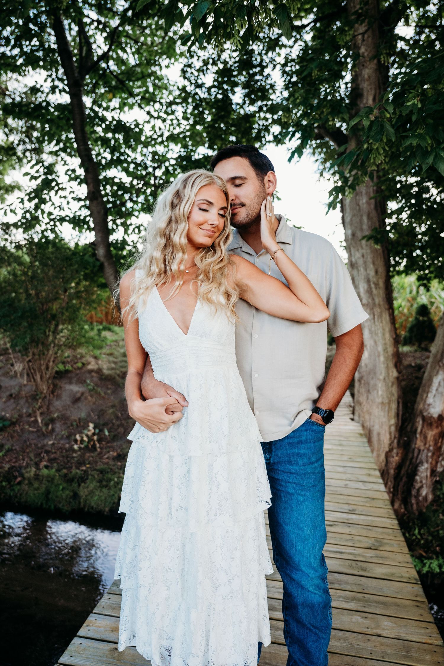 A couple shares romantic moments on a wooden boardwalk surrounded by trees and water on a sunny day.