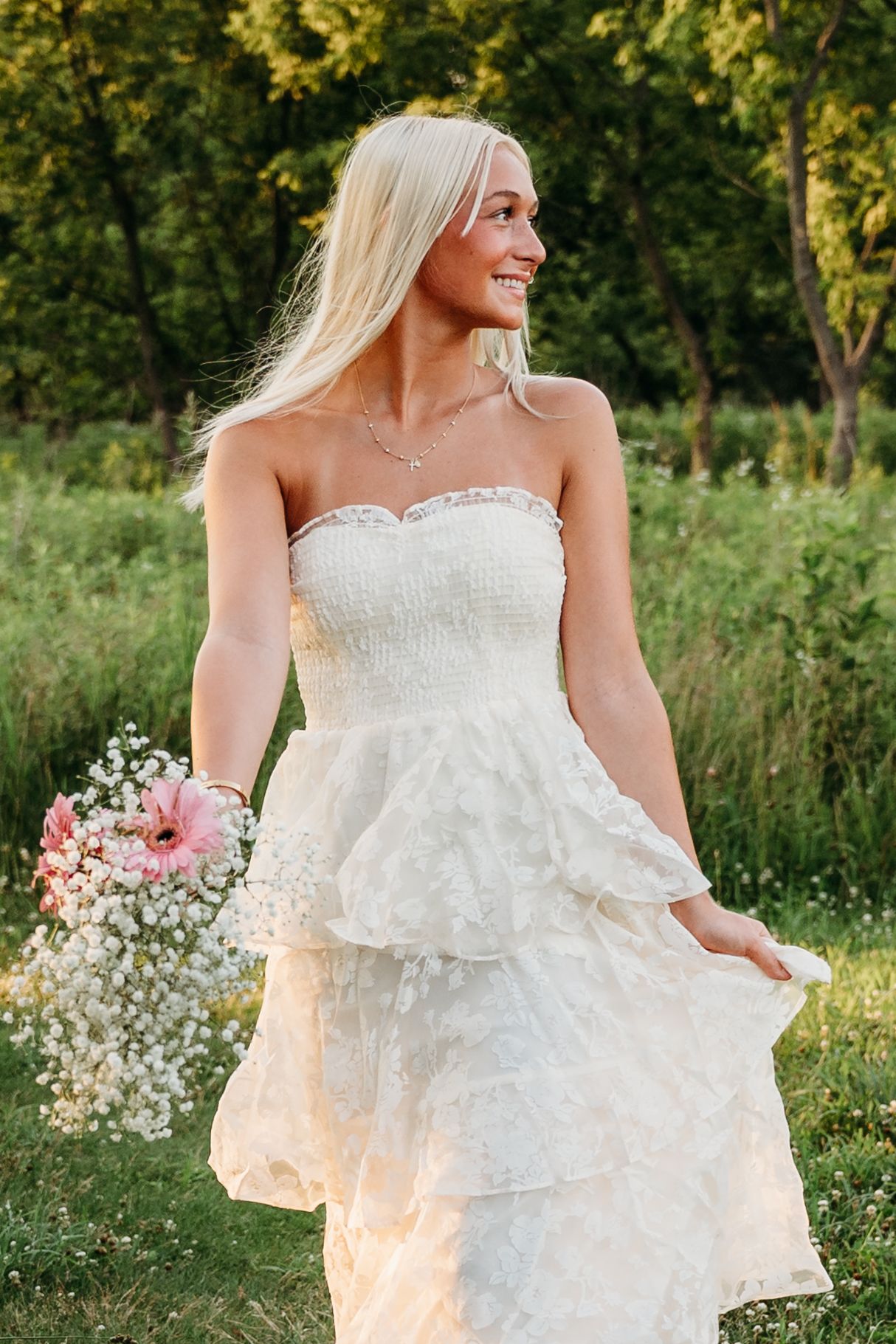 A person in a white lace wedding dress holding a pink and white bouquet poses in a sunny outdoor garden setting.