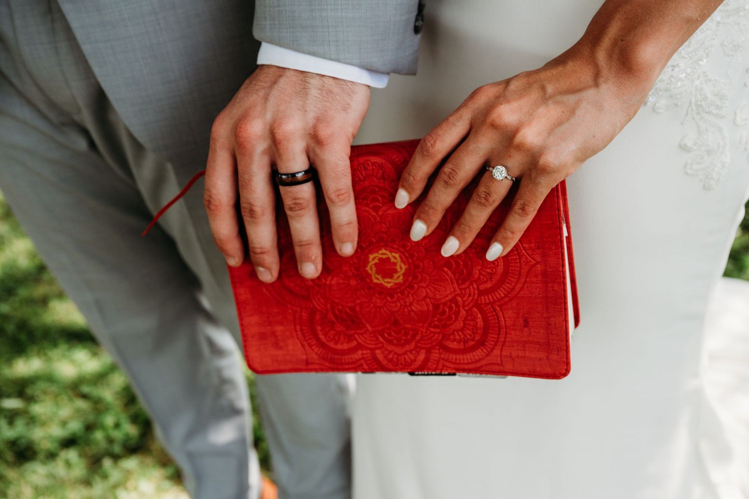 Two hands wearing wedding rings hold a decorative red clutch purse with ornate embroidery.