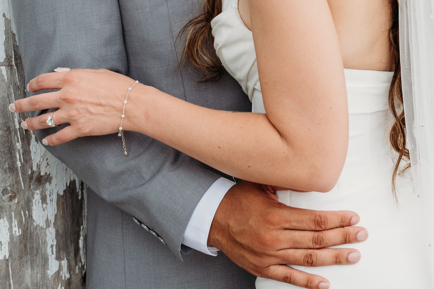 Close-up detail of bride and groom embracing, showing wedding rings and formal attire in gray and white.