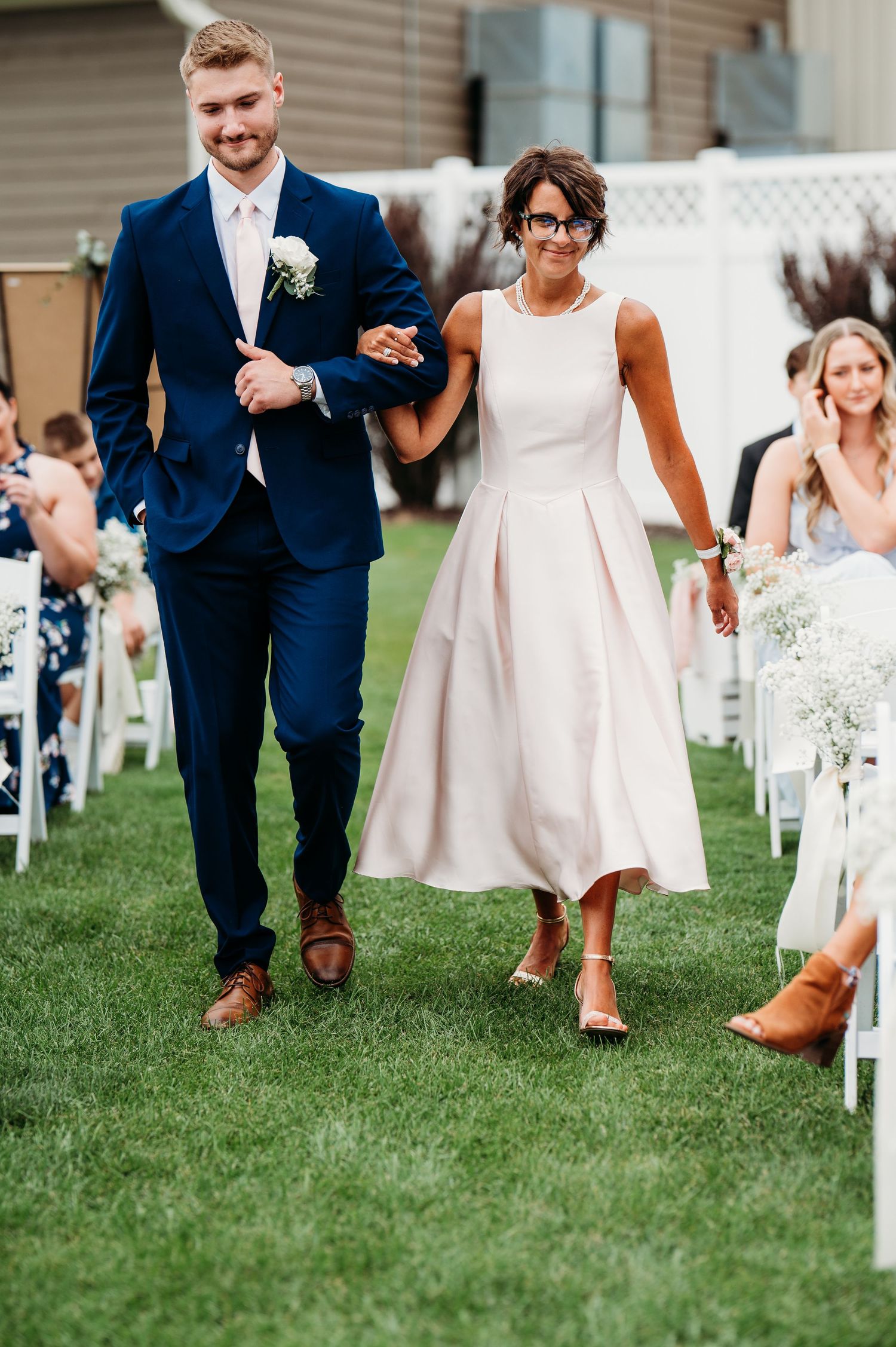 A couple walks arm in arm down a grassy aisle at an outdoor wedding ceremony wearing navy and cream formal attire.