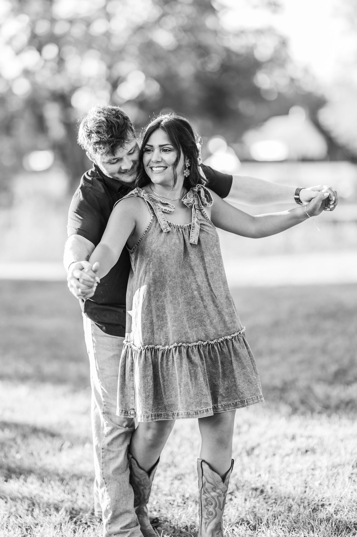 A couple playfully dances together in a grassy field on a sunny day, captured in black and white.
