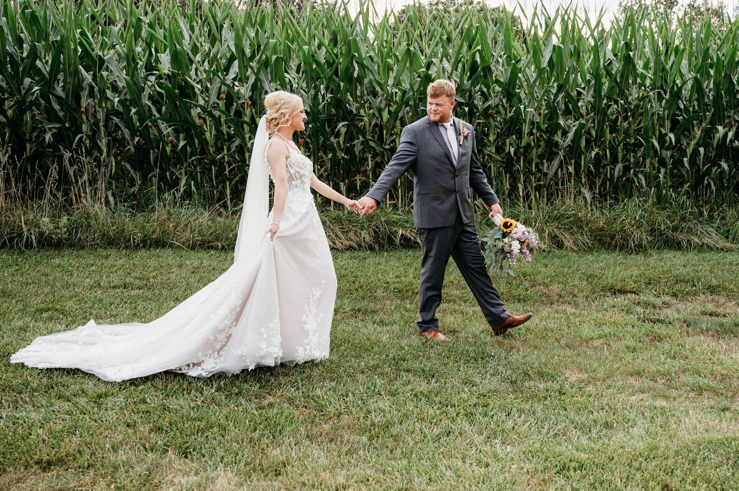 A couple walks hand in hand near a tall cornfield at their rustic outdoor wedding.