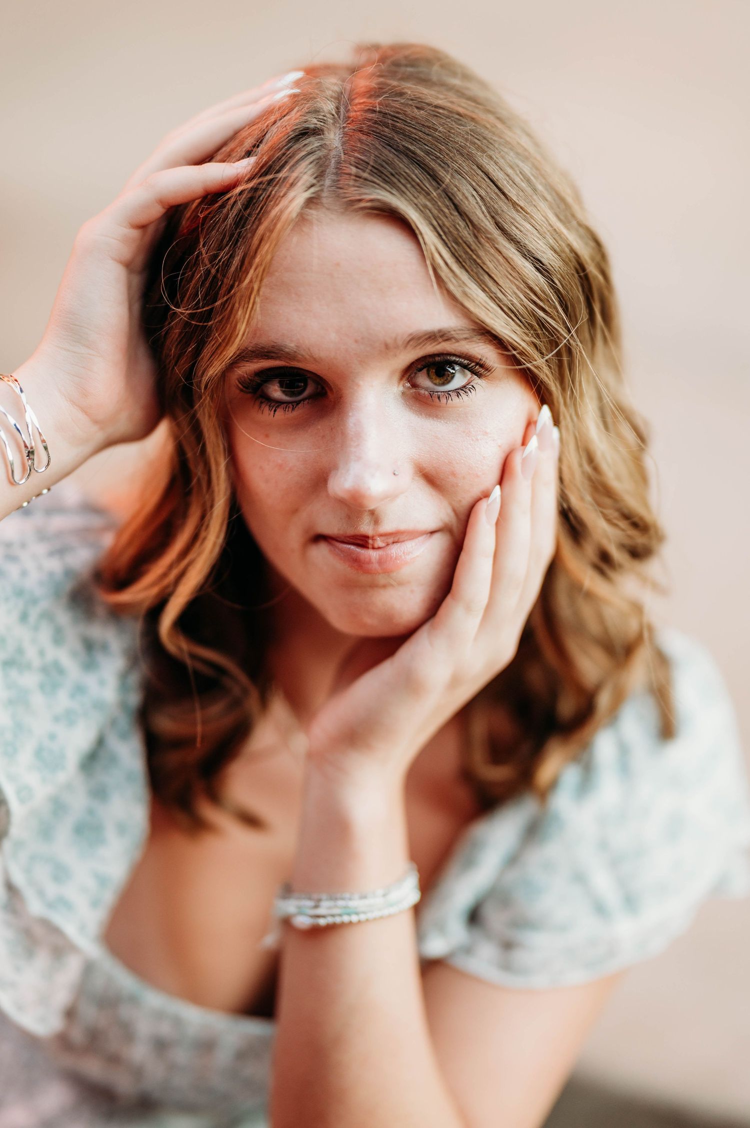 Close-up portrait of young woman with wavy hair resting face in hands wearing bracelets indoors.