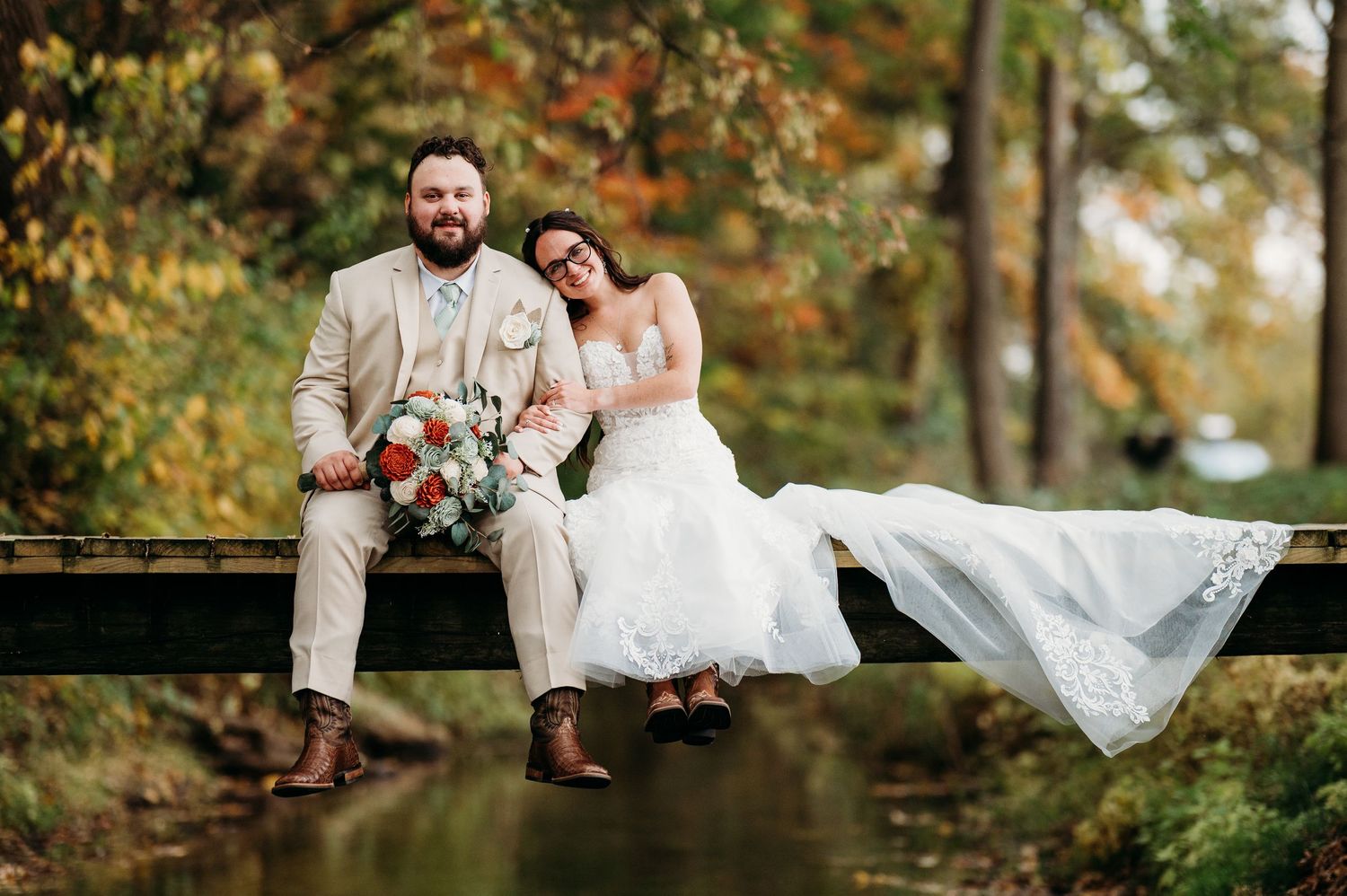 A bride and groom sit together on a wooden bridge in an autumn forest setting with her dress flowing down.