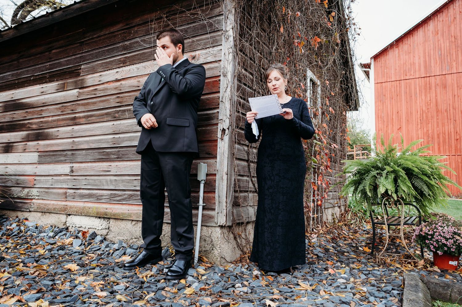 Two people in formal black attire stand against a rustic wooden wall with a red barn in the background.