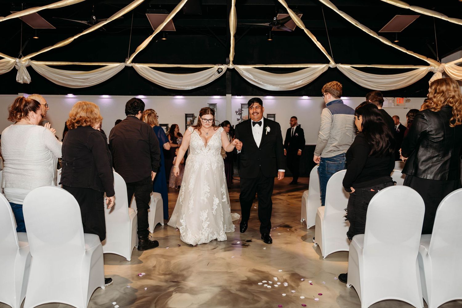 Wedding guests dance and celebrate on a wooden dance floor at an indoor reception venue with white chairs and draping.