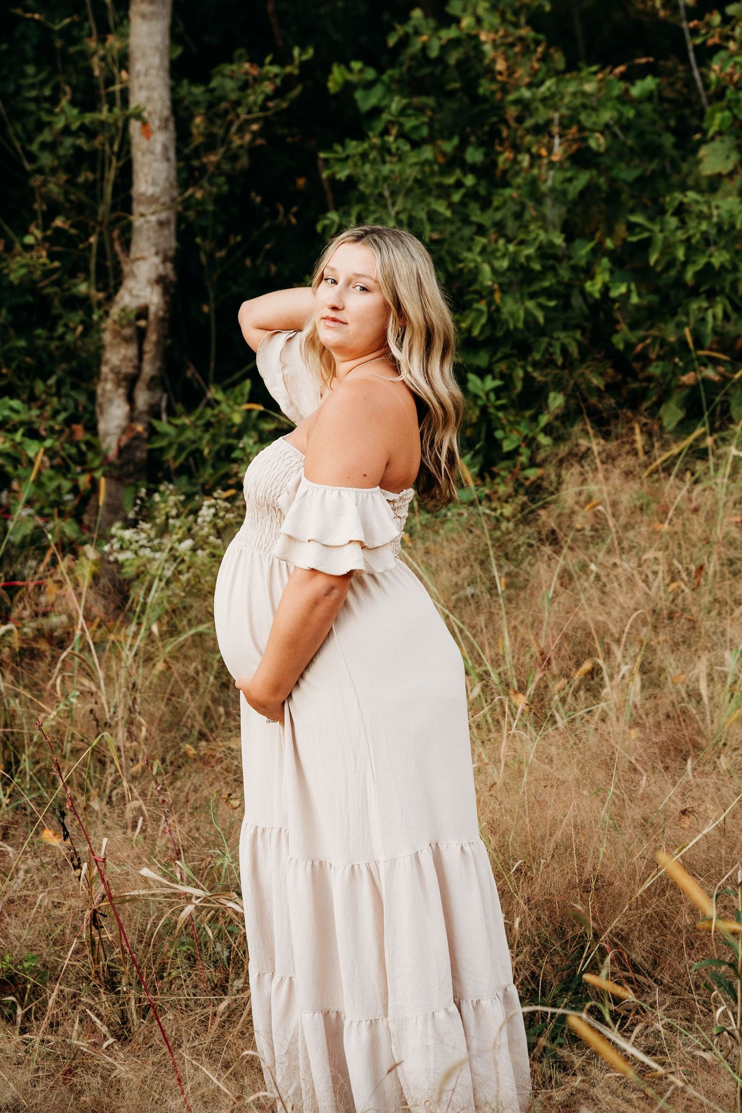 A maternity portrait showing a white off-shoulder maxi dress against a natural outdoor backdrop with tall grass.