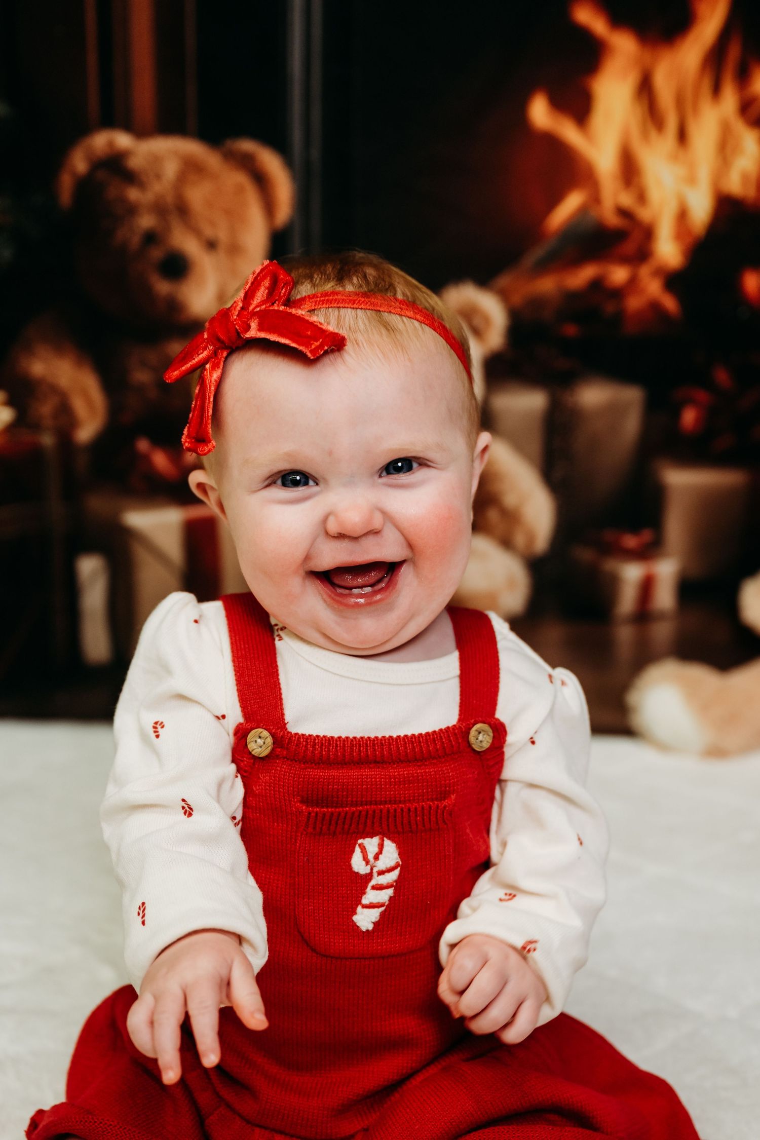 A baby wearing red overalls and white shirt smiles while sitting by a fireplace during Christmas.