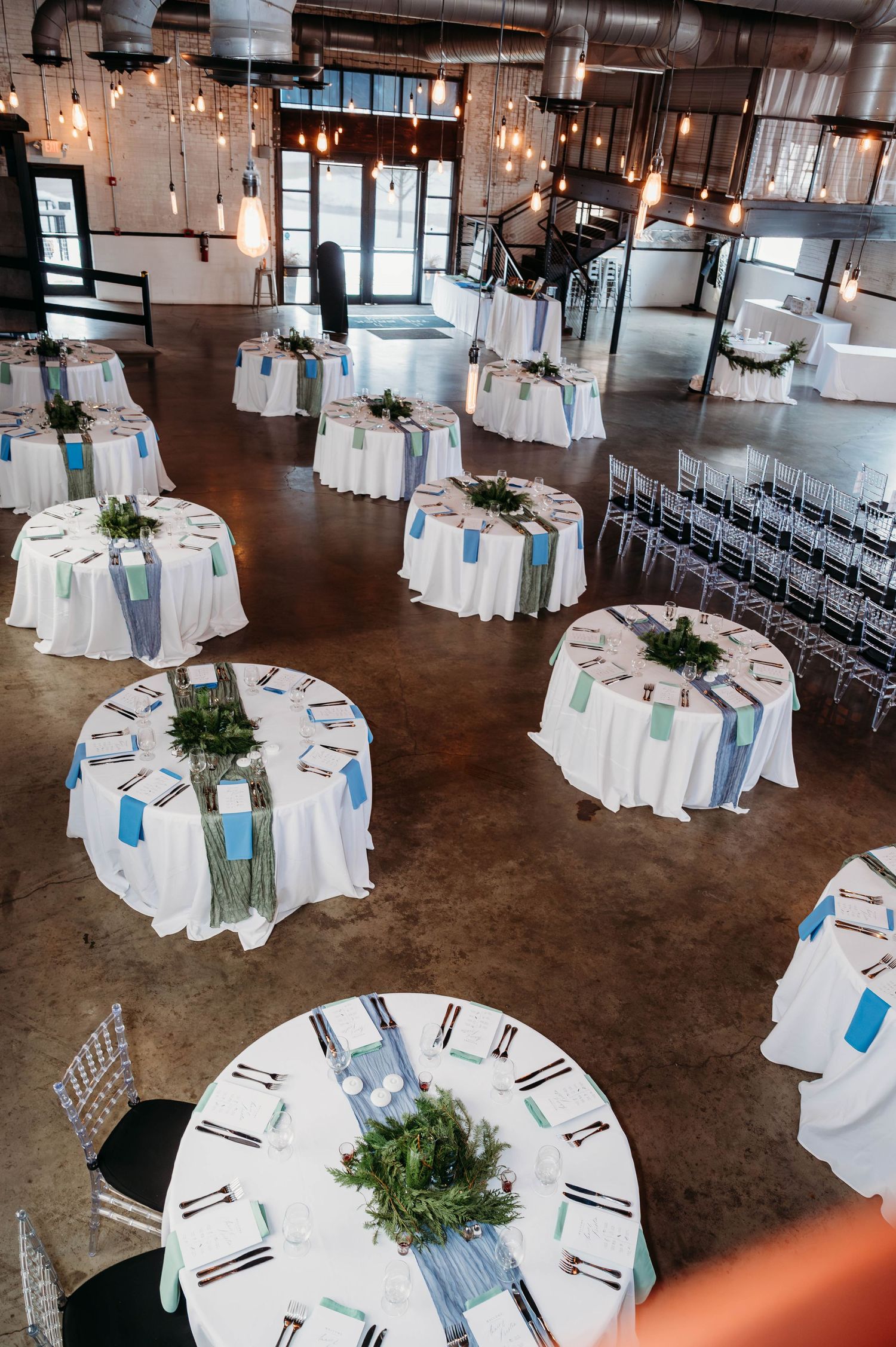 Wedding reception venue setup with round tables, white linens, and hanging lights creating an elegant industrial atmosphere.