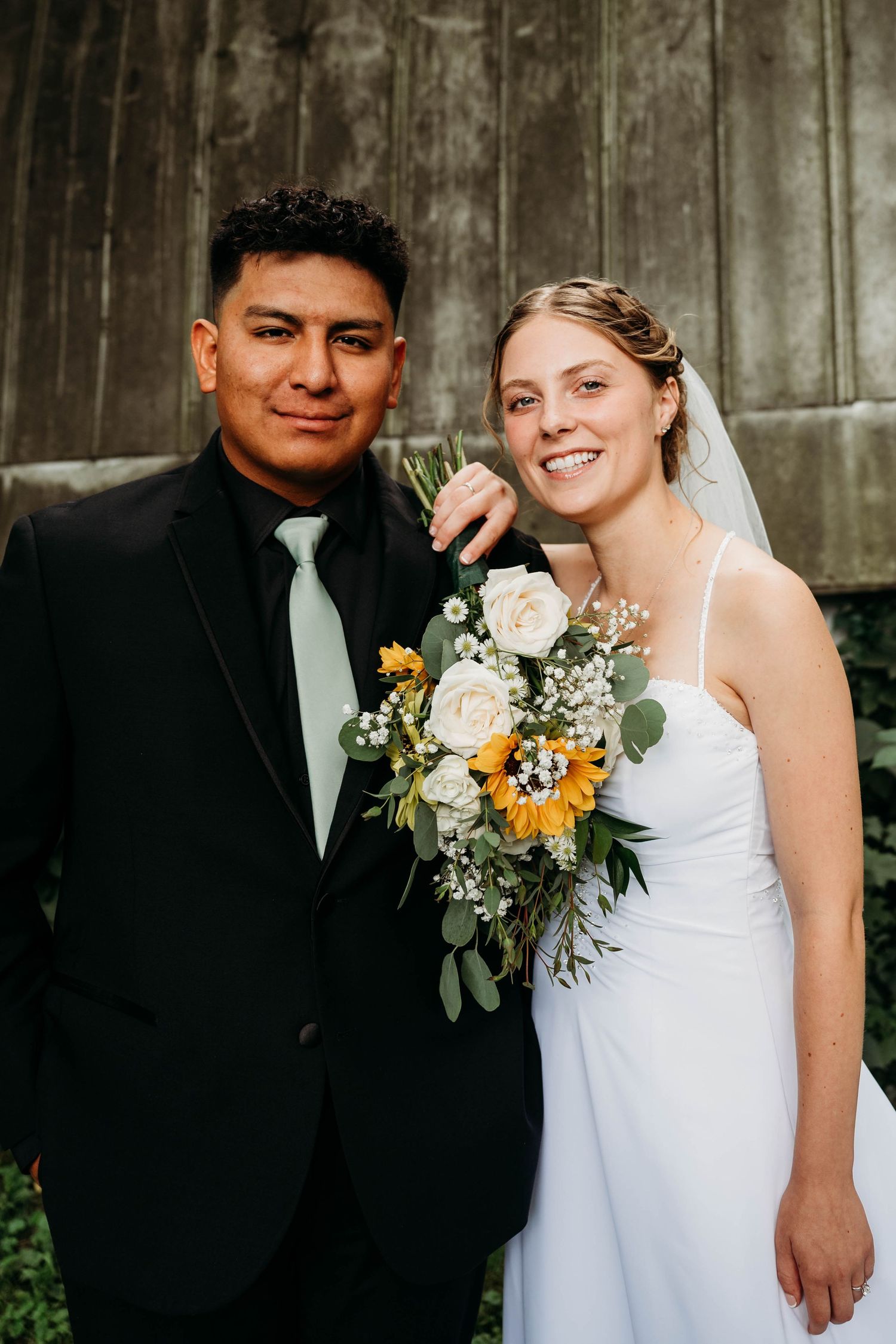 Bride and groom posing together, bride holding sunflower and white rose bouquet, groom in black suit with mint tie.