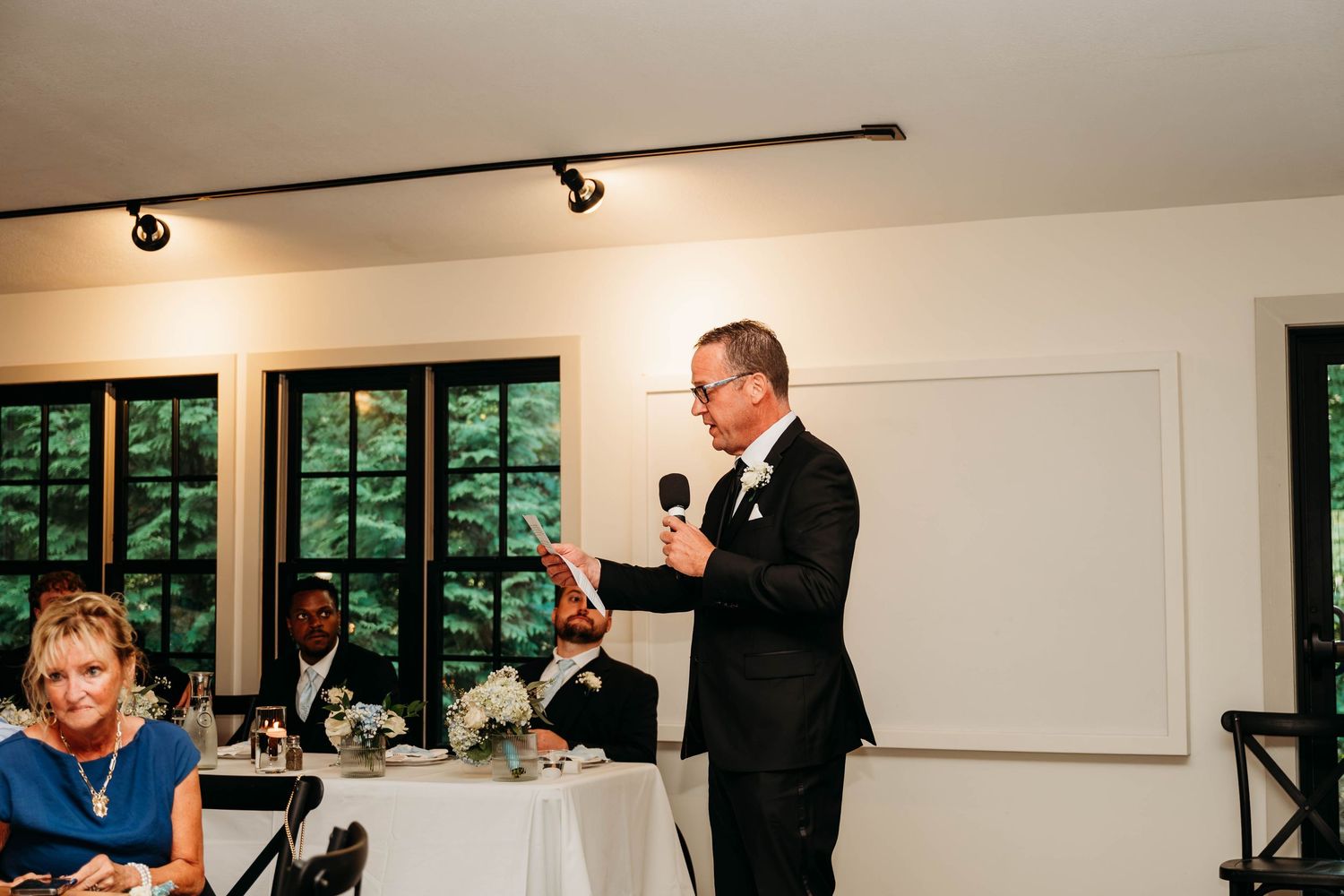 Man in black suit giving a speech with microphone at an elegant wedding reception indoors.