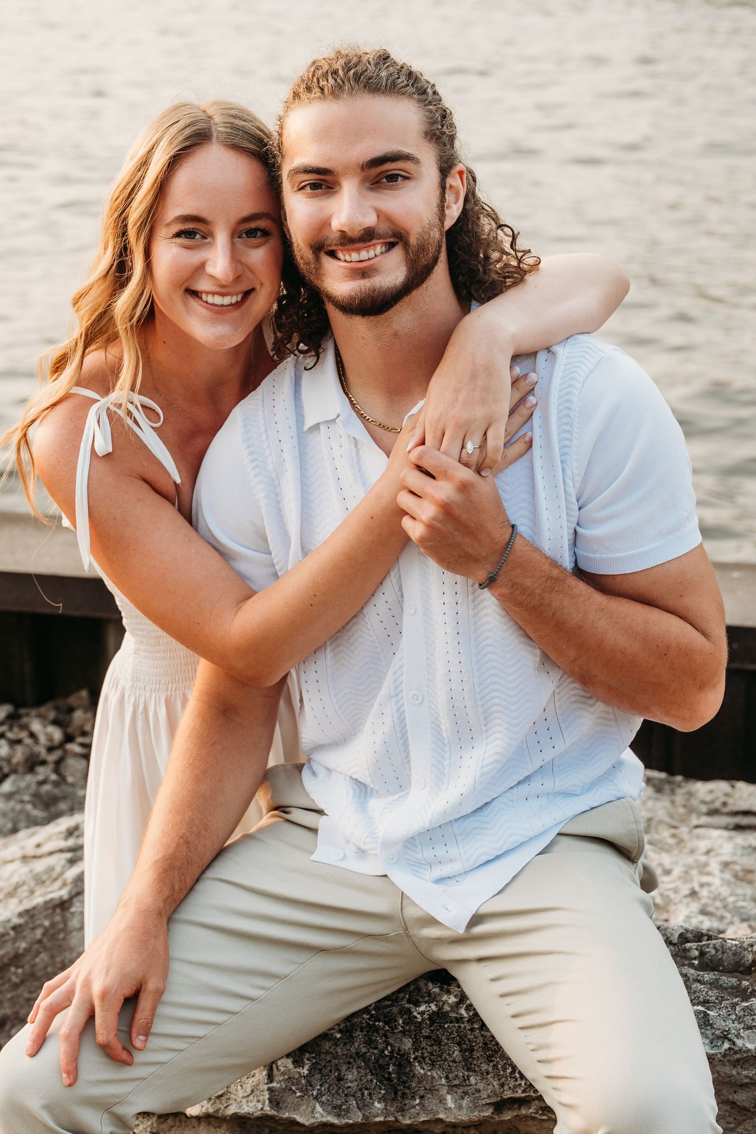 Smiling couple embracing on rocky waterfront, woman in white dress hugging man in light blue polo shirt outdoors.
