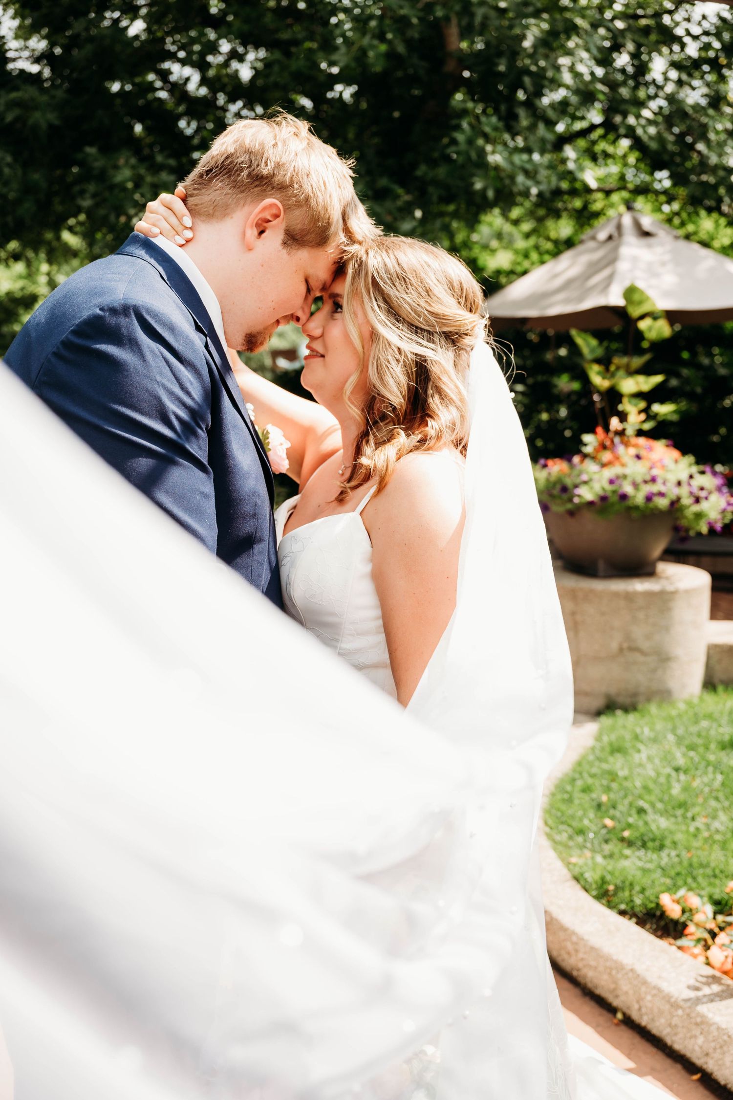 A bride and groom share a romantic moment in a garden, her veil flowing in the breeze, as they touch foreheads tenderly.