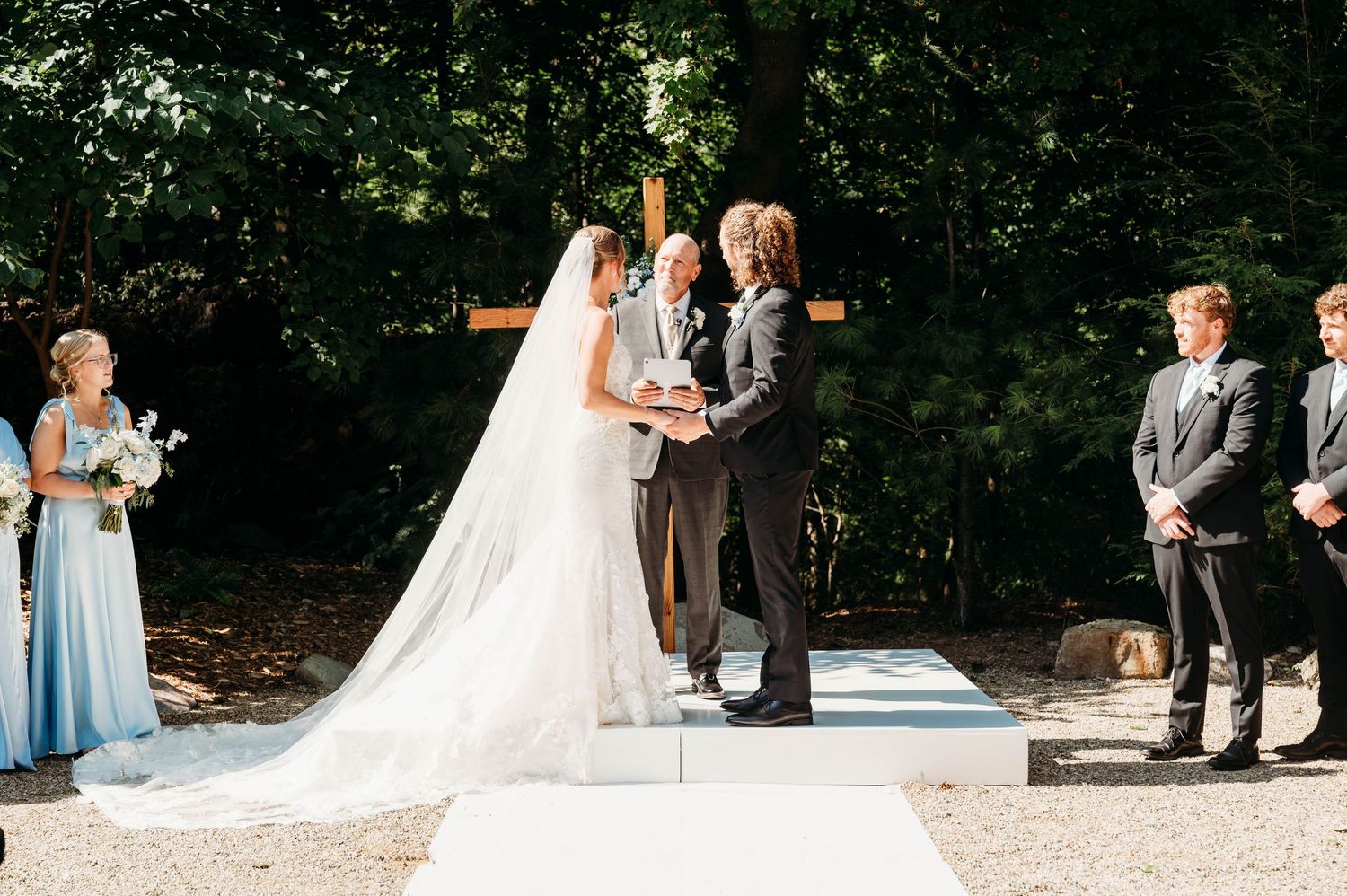 Outdoor wedding ceremony with bride in white gown and veil, groom, and officiant at wooden cross altar.