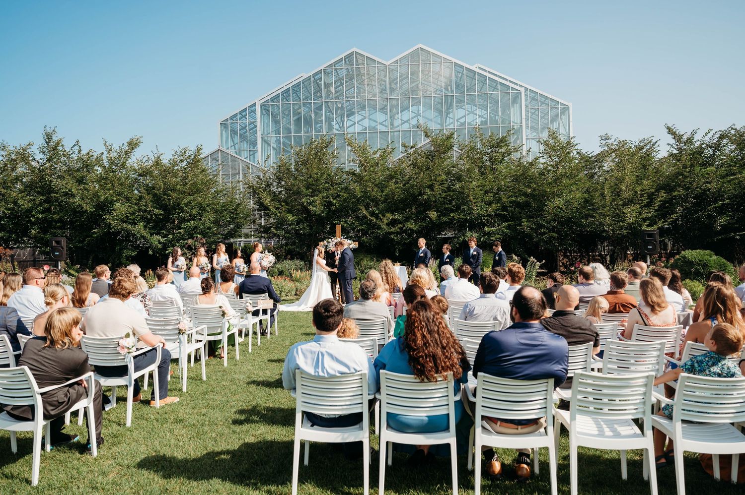 Outdoor wedding ceremony with guests seated in white chairs on a lawn, facing couple at altar near a glass greenhouse.