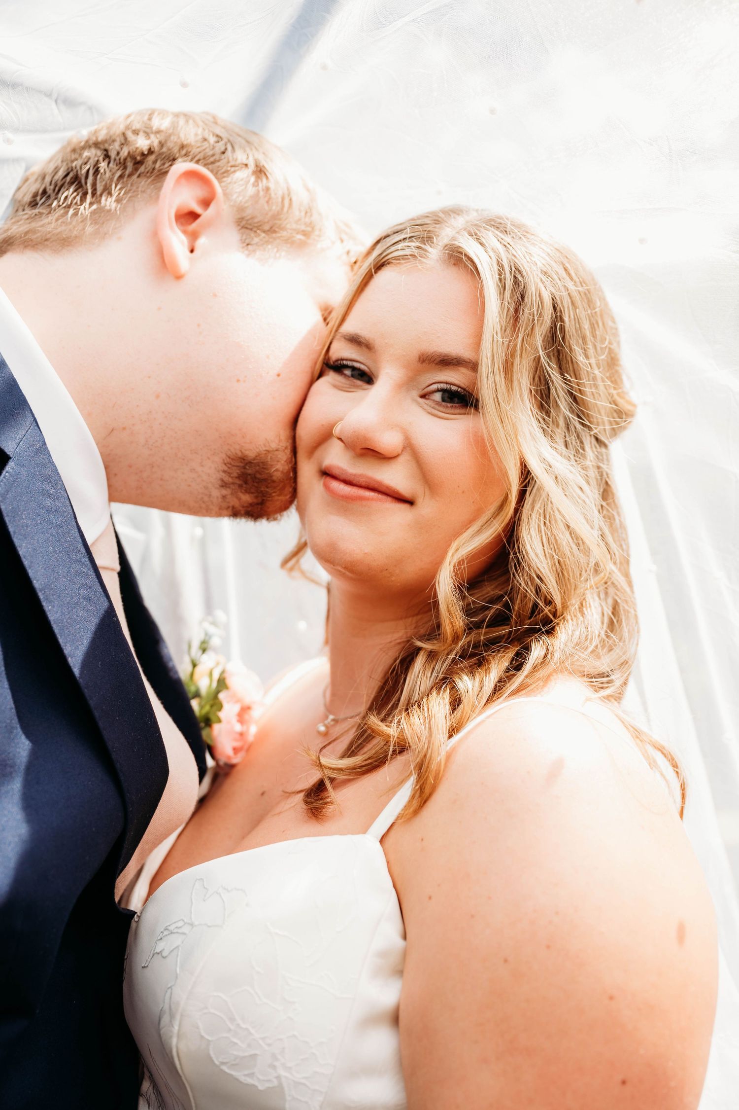 Groom kissing bride on cheek under veil, bride smiling at camera in white dress, romantic wedding portrait.