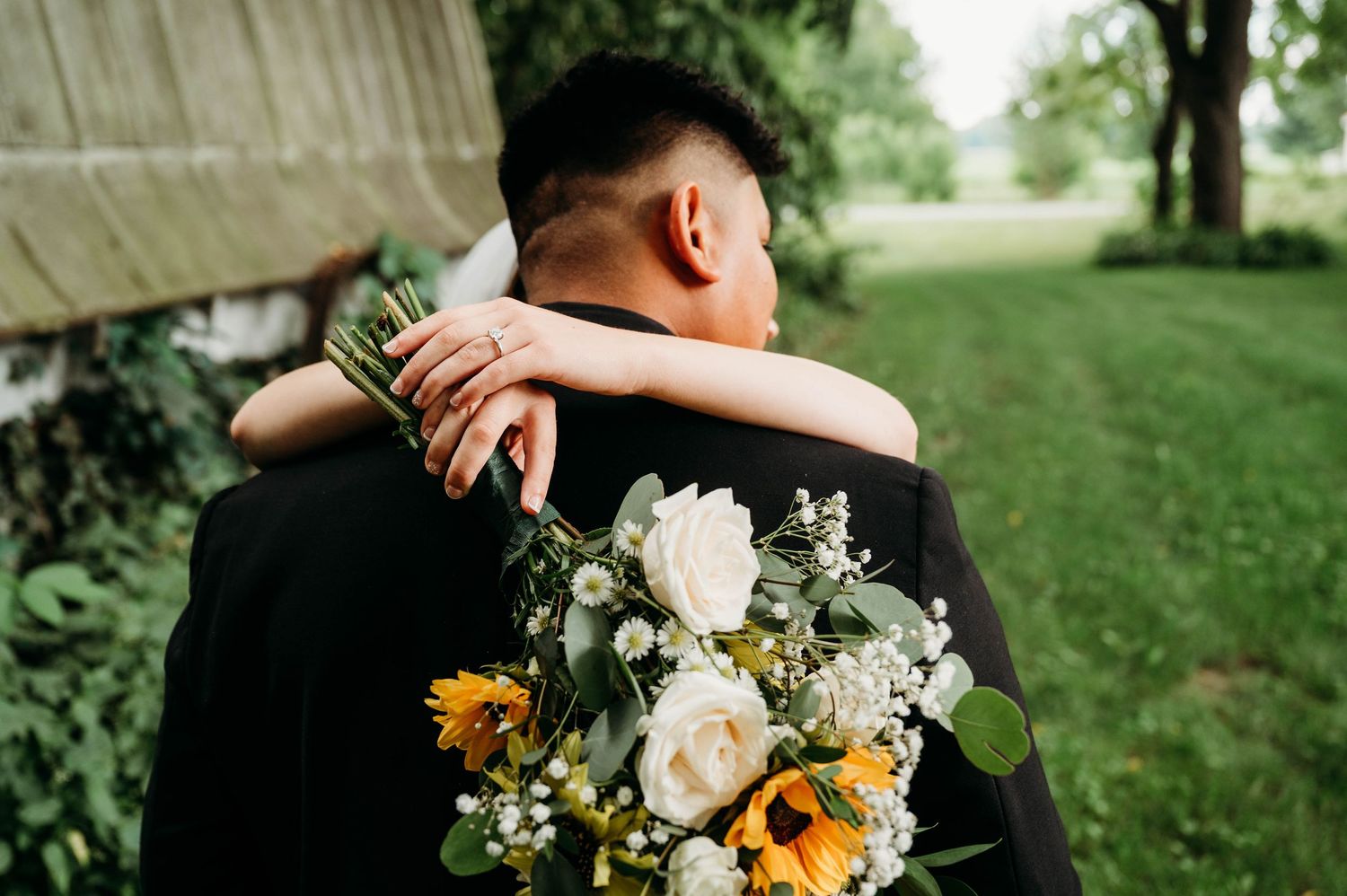Couple embracing at wedding, bride holding sunflower and white rose bouquet with greenery, groom in black suit outdoors.