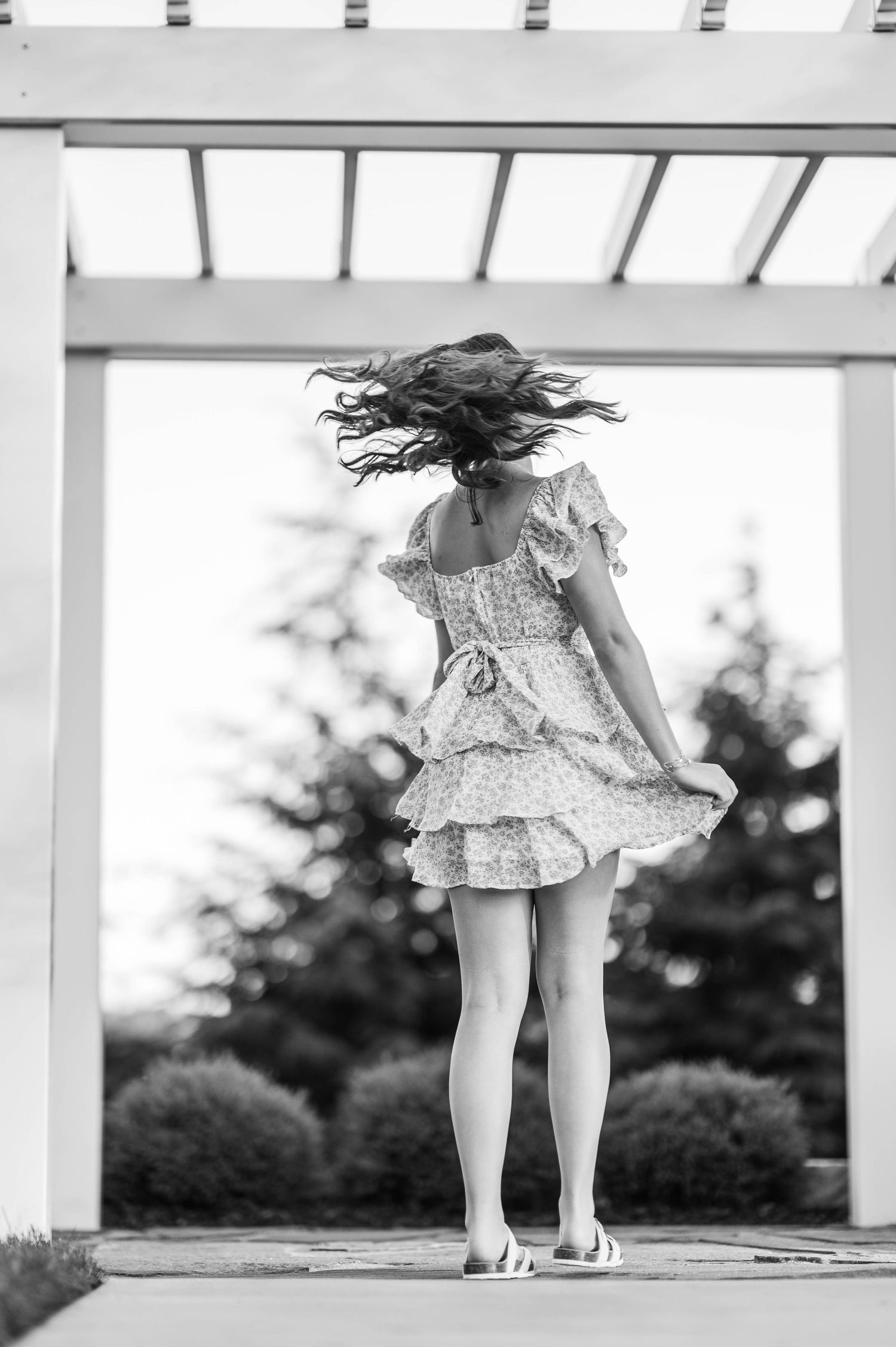 Black and white photo of a girl in a floral dress twirling under a white pergola in a garden.