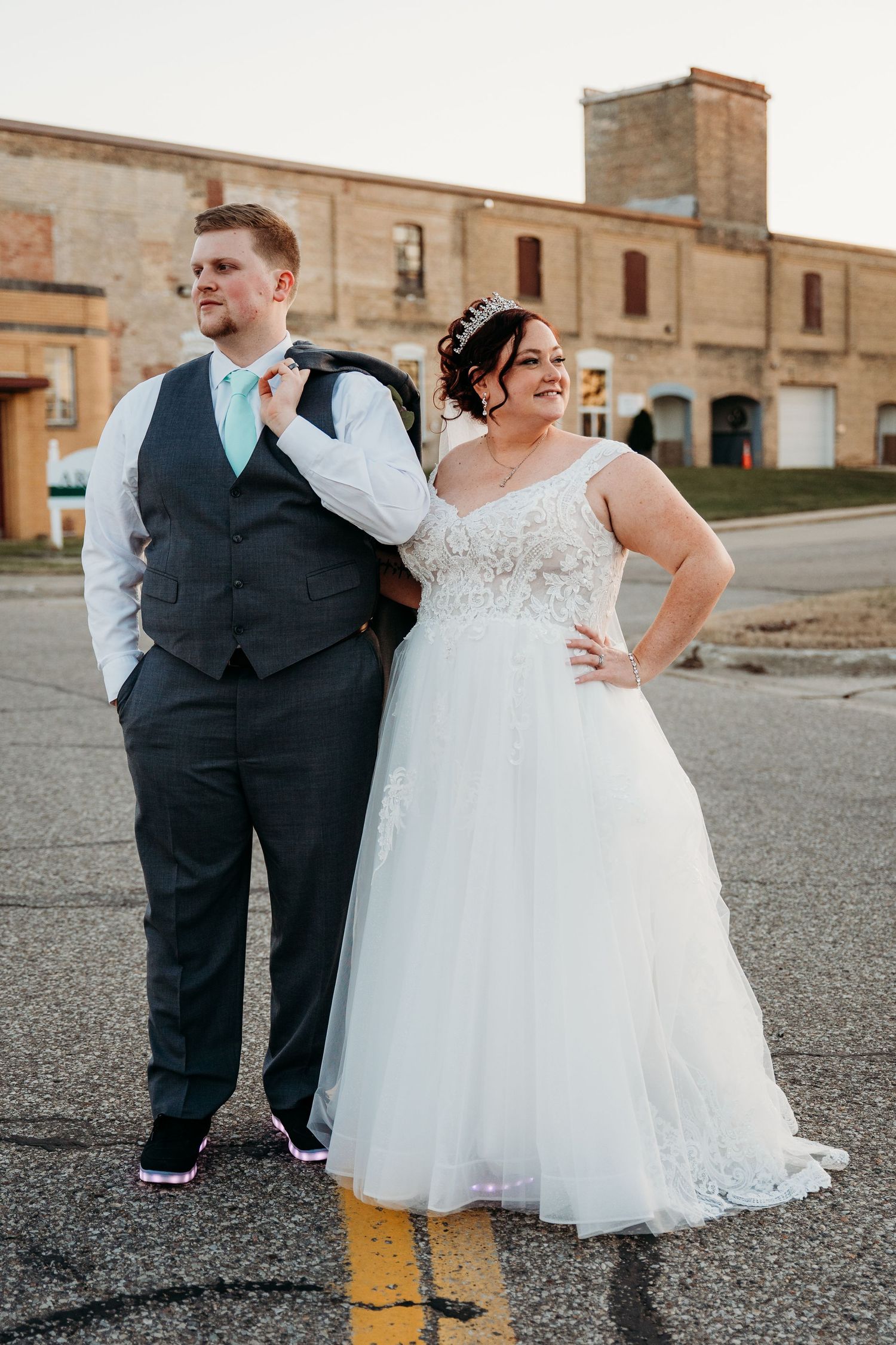 A newlywed couple poses casually on a street, with the bride in a lace gown and groom in vest and tie.
