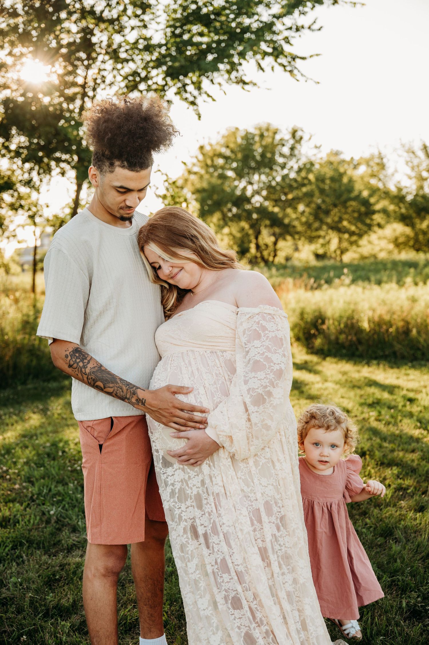 A family poses together in a sunlit field with a pregnant woman in a white lace dress and coordinating dusty rose outfits.