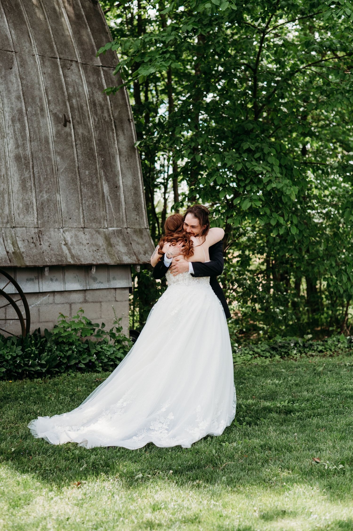 Two people embrace while wearing elegant formal attire outdoors near a stone wall and green trees.