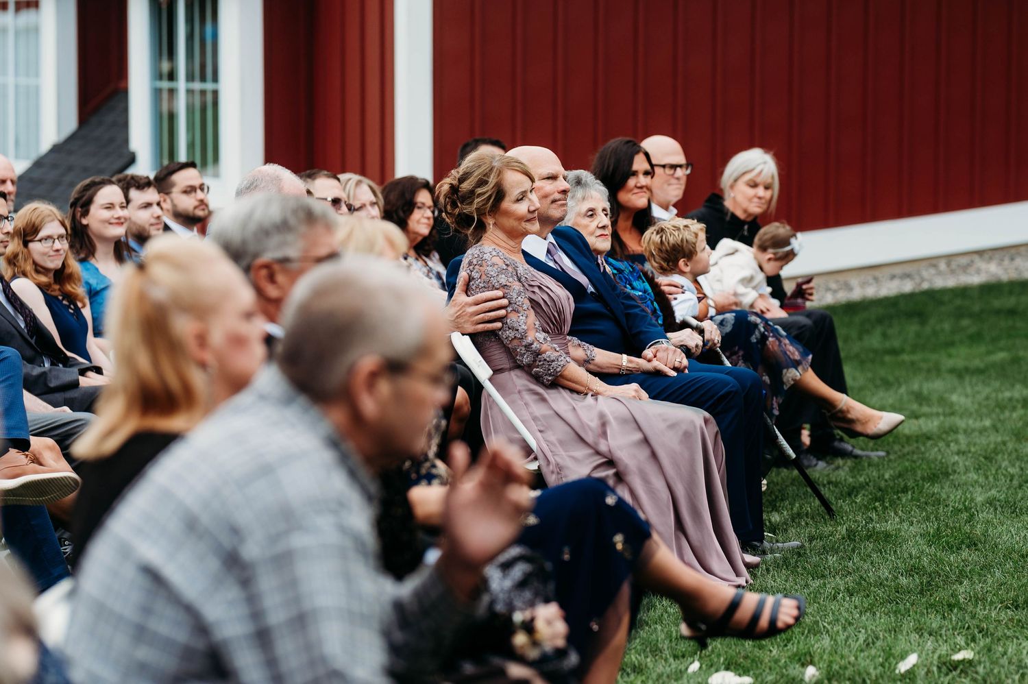 Wedding guests seated on chairs on a lawn watching an outdoor ceremony next to a red barn building.