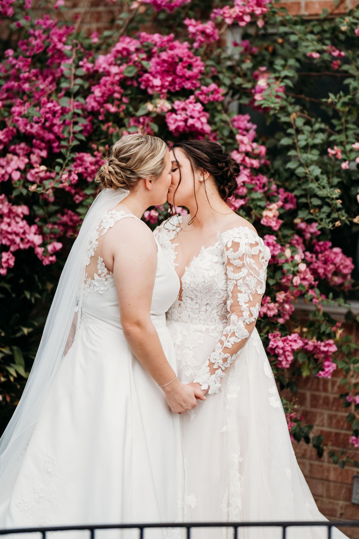 Two people in white wedding dresses standing together among vibrant pink bougainvillea flowers and brick walls.