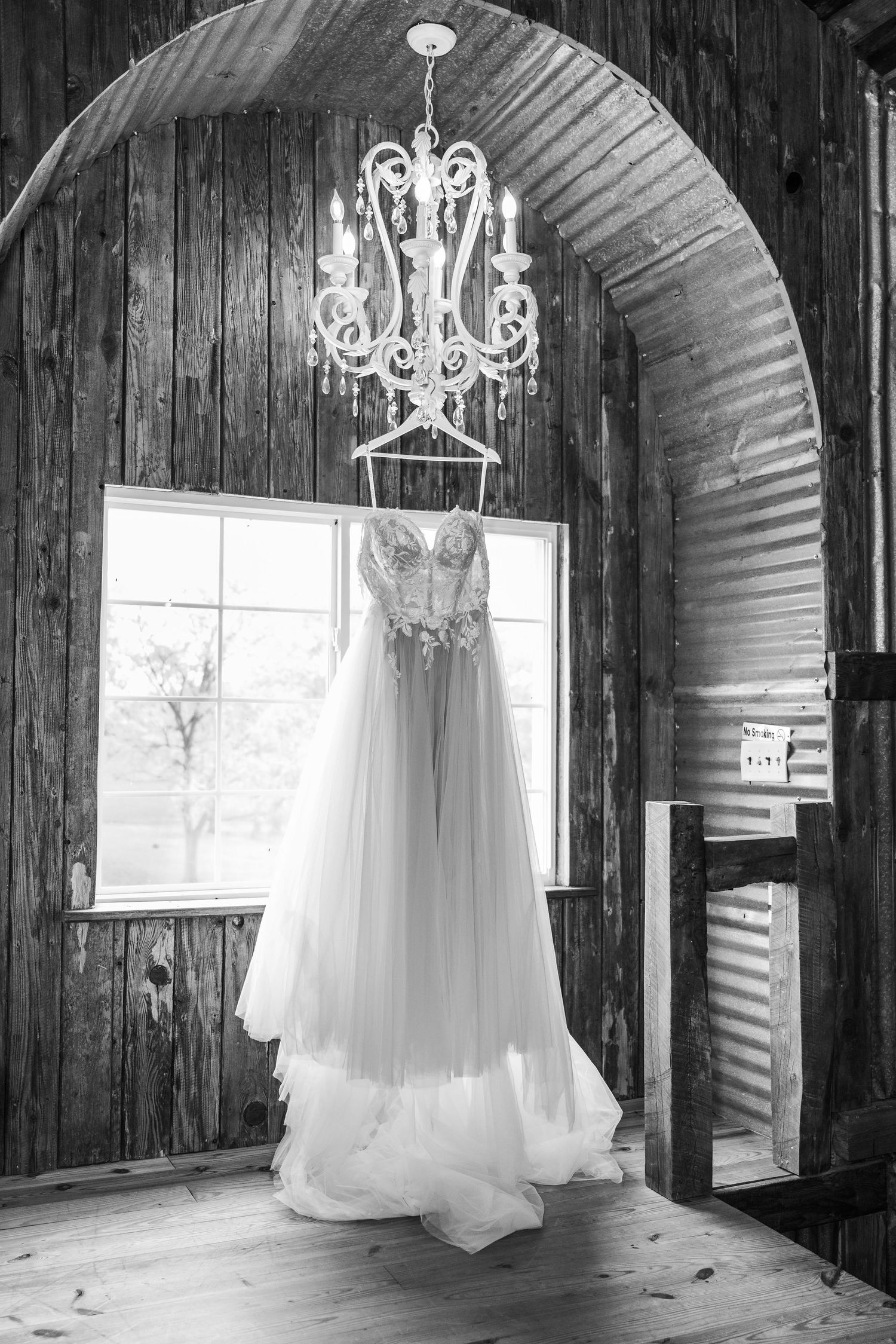 White wedding dress hangs elegantly in rustic barn venue with crystal chandeliers and natural light streaming through windows.