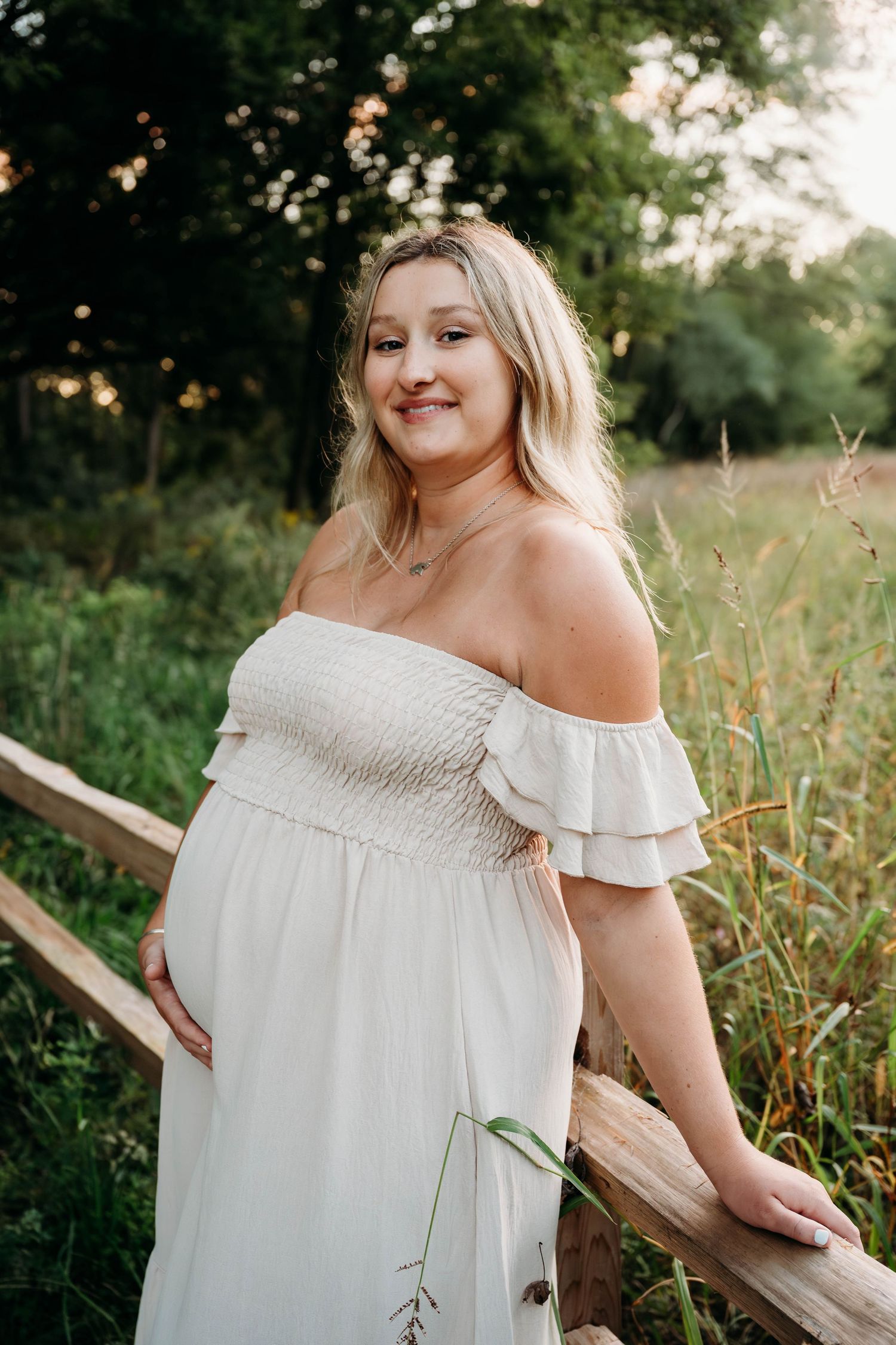 A person in a white off-shoulder smocked dress poses outdoors in a natural setting with greenery and sunlight.