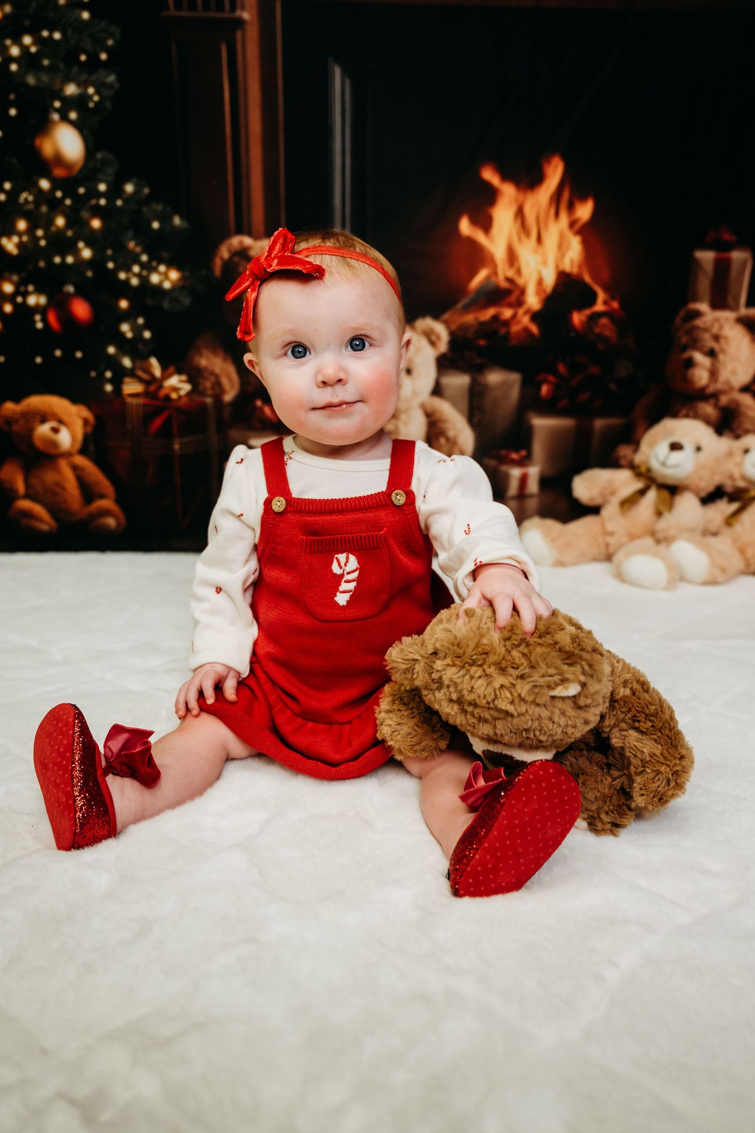 Festive holiday scene with red corduroy overalls, teddy bears, and a cozy fireplace with Christmas tree in background.
