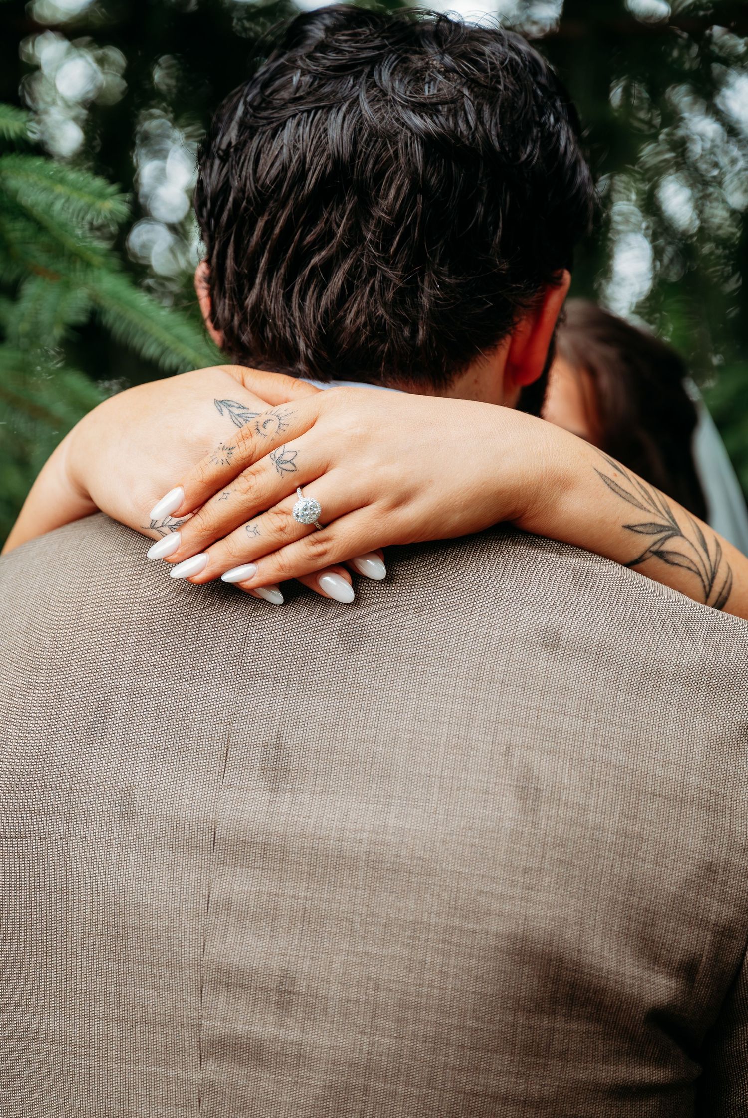 Bride wraps her tattooed hand with manicured white nails around groom in tan suit during outdoor wedding.