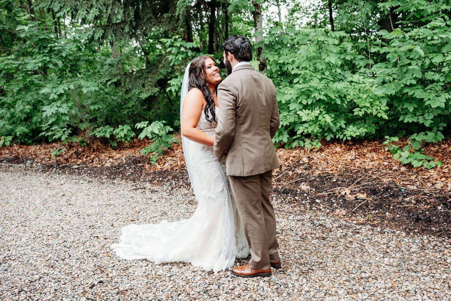 Bride and groom share a romantic moment surrounded by lush green trees on a gravel path at an outdoor wedding.