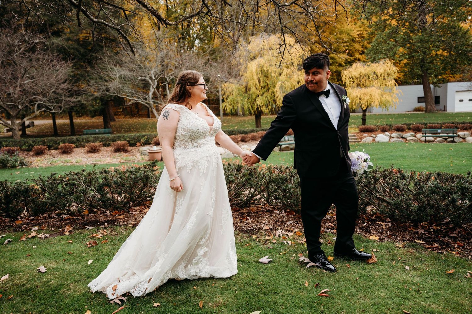 A couple in wedding attire holds hands while walking through a garden with autumn foliage in the background.