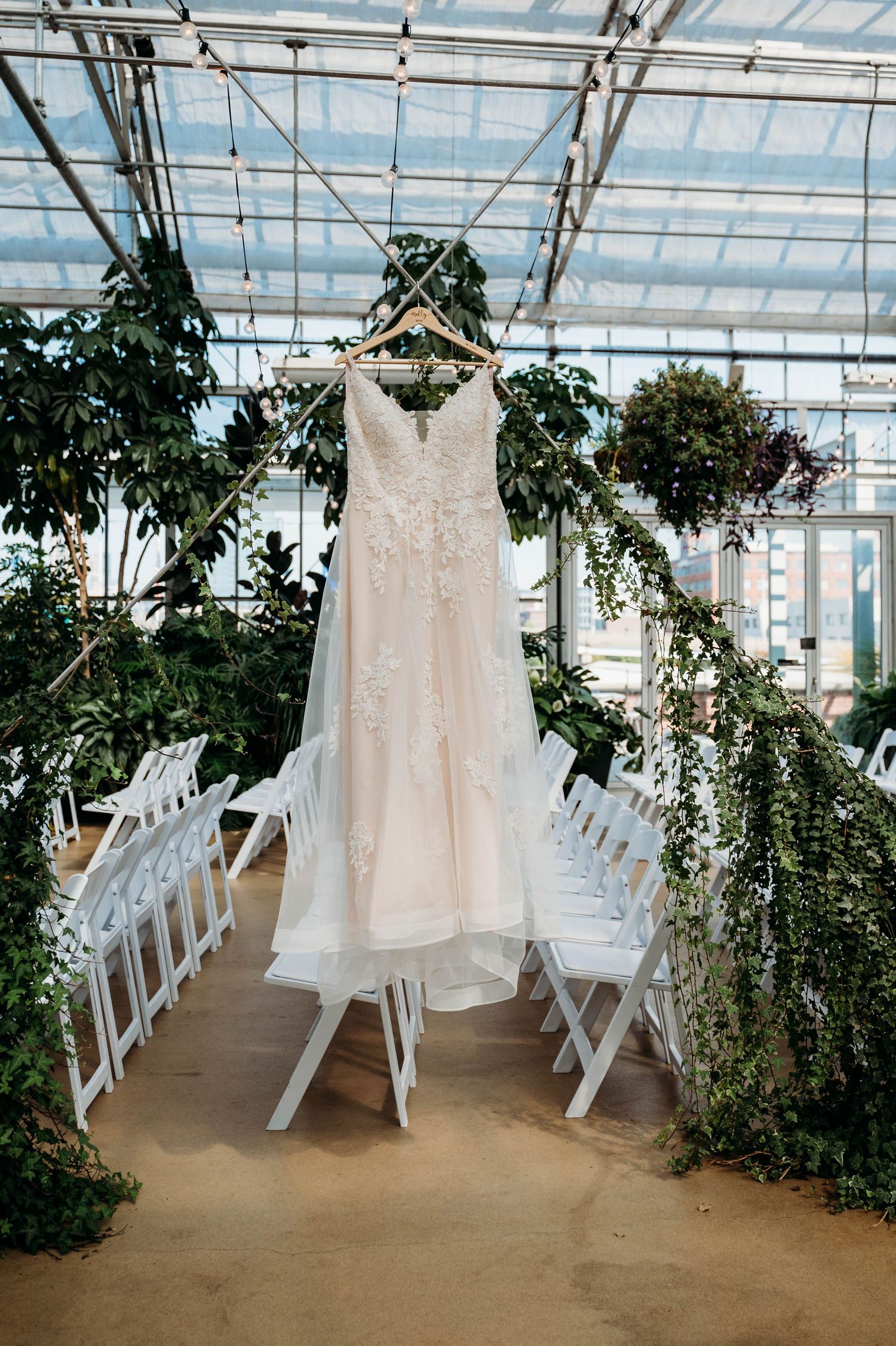 A white lace wedding dress hangs in a greenhouse venue with white folding chairs and lush greenery surrounding it.