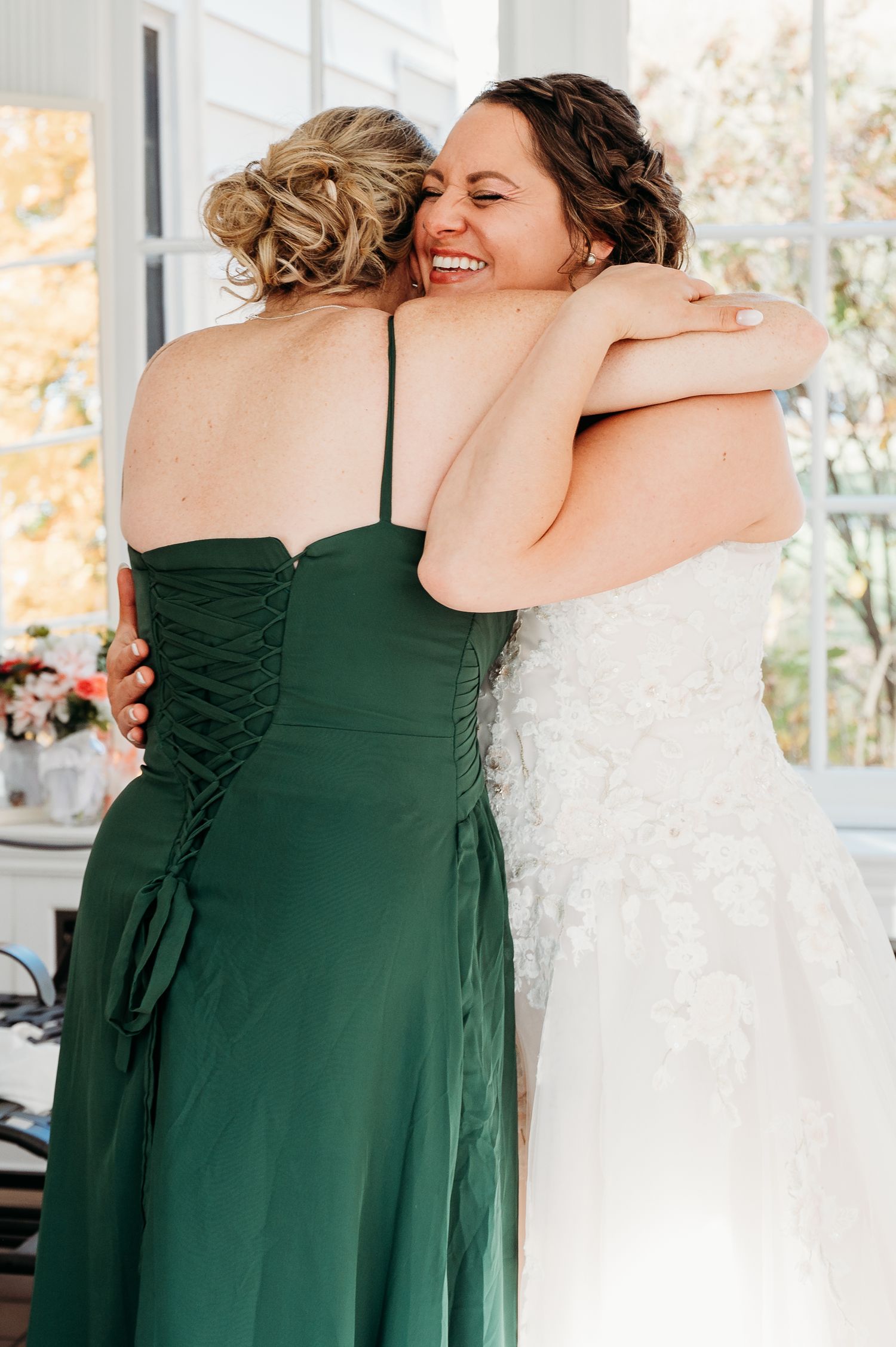 A joyful moment shared between someone in a forest green bridesmaid dress and a bride in a white lace gown.