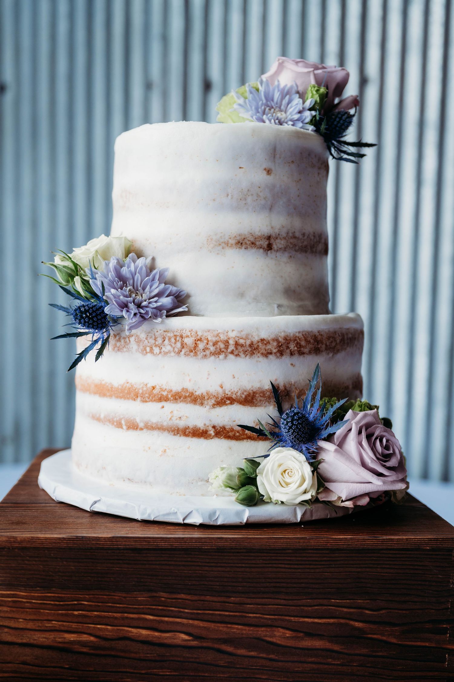 Two-tiered semi-naked wedding cake decorated with blue thistles and pink roses on wooden stand.