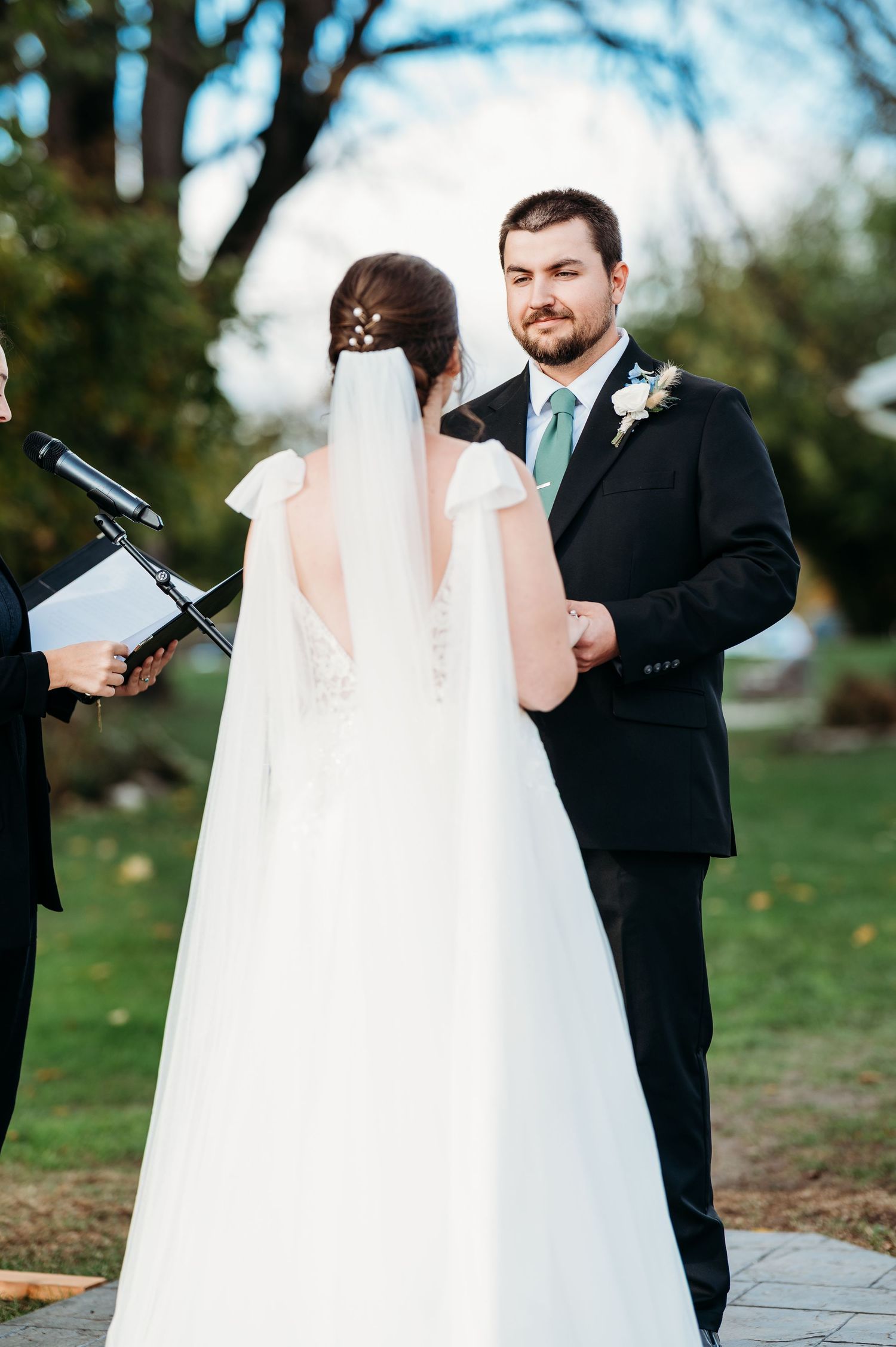 Groom in black suit with green tie smiling at bride in white gown and veil during outdoor wedding ceremony.
