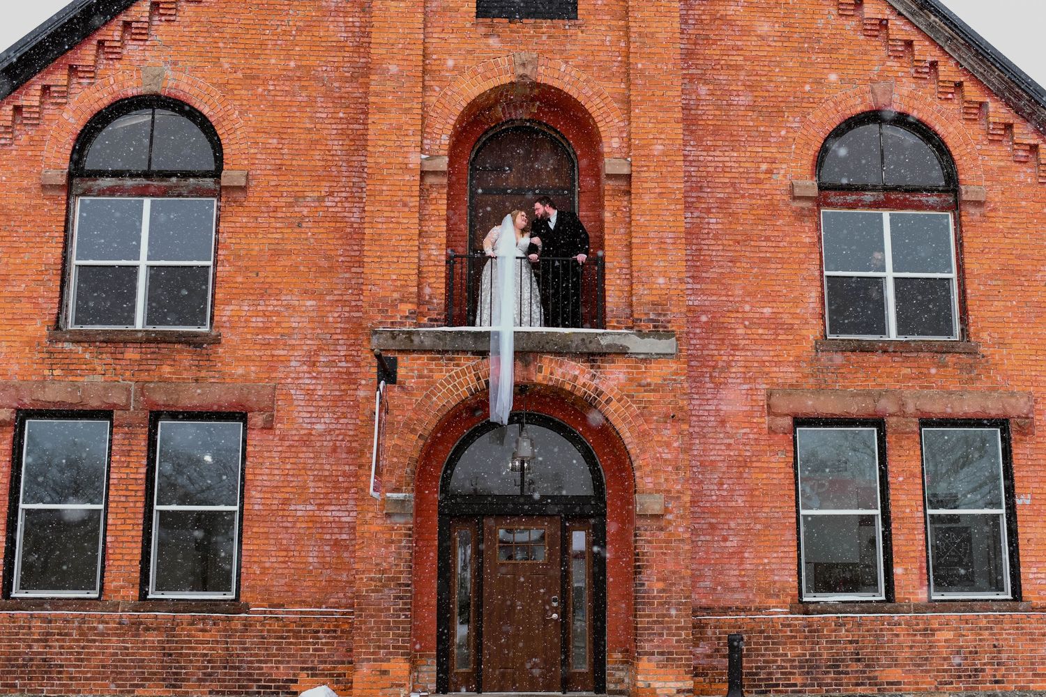 Gothic-style red brick church building with arched windows and a person in white dress standing on balcony entrance.
